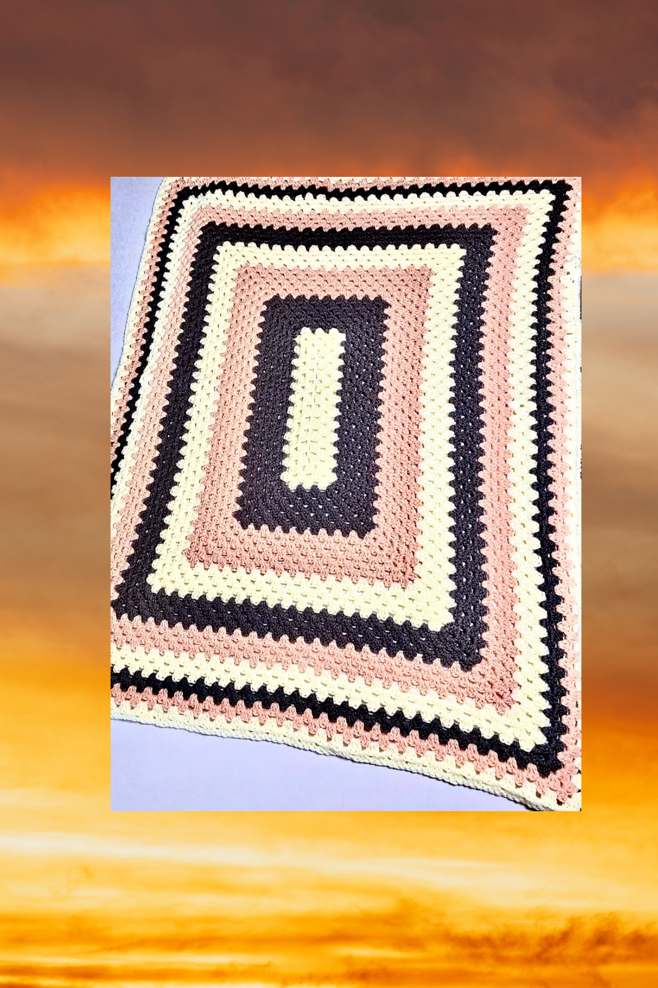 A crocheted rug with a geometric pattern of black, pink, and cream-colored squares layered inside each other, placed on a flat surface