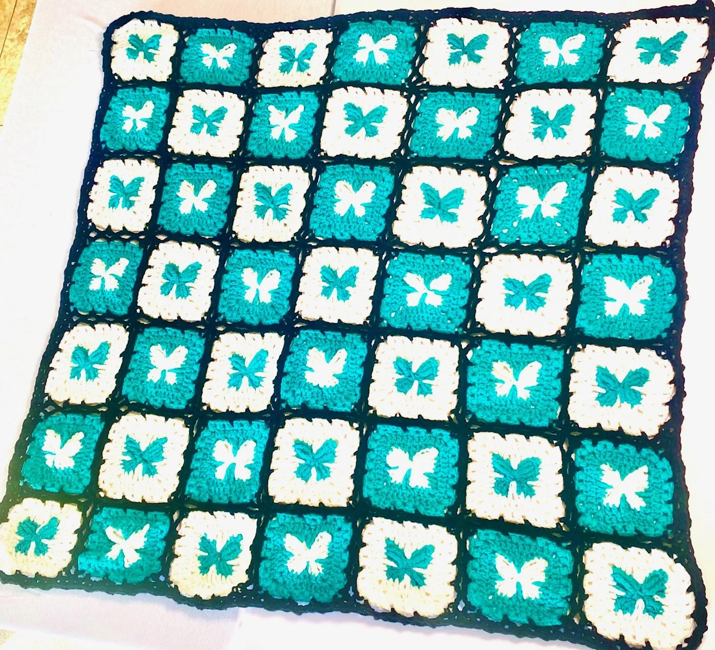 A crocheted blanket with a pattern of white and turquoise squares, each featuring a butterfly design, bordered in black.