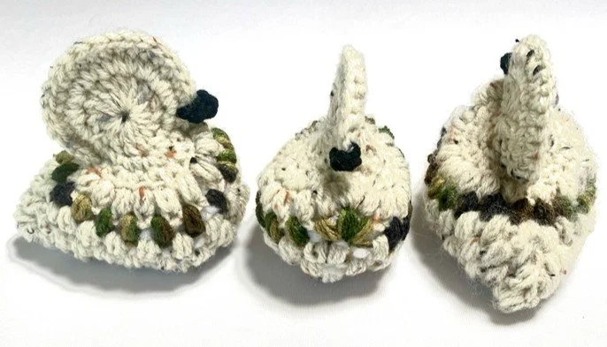 Three crocheted swan figurines in white with black beaks, green accents, and dark feathers, placed on a white background.