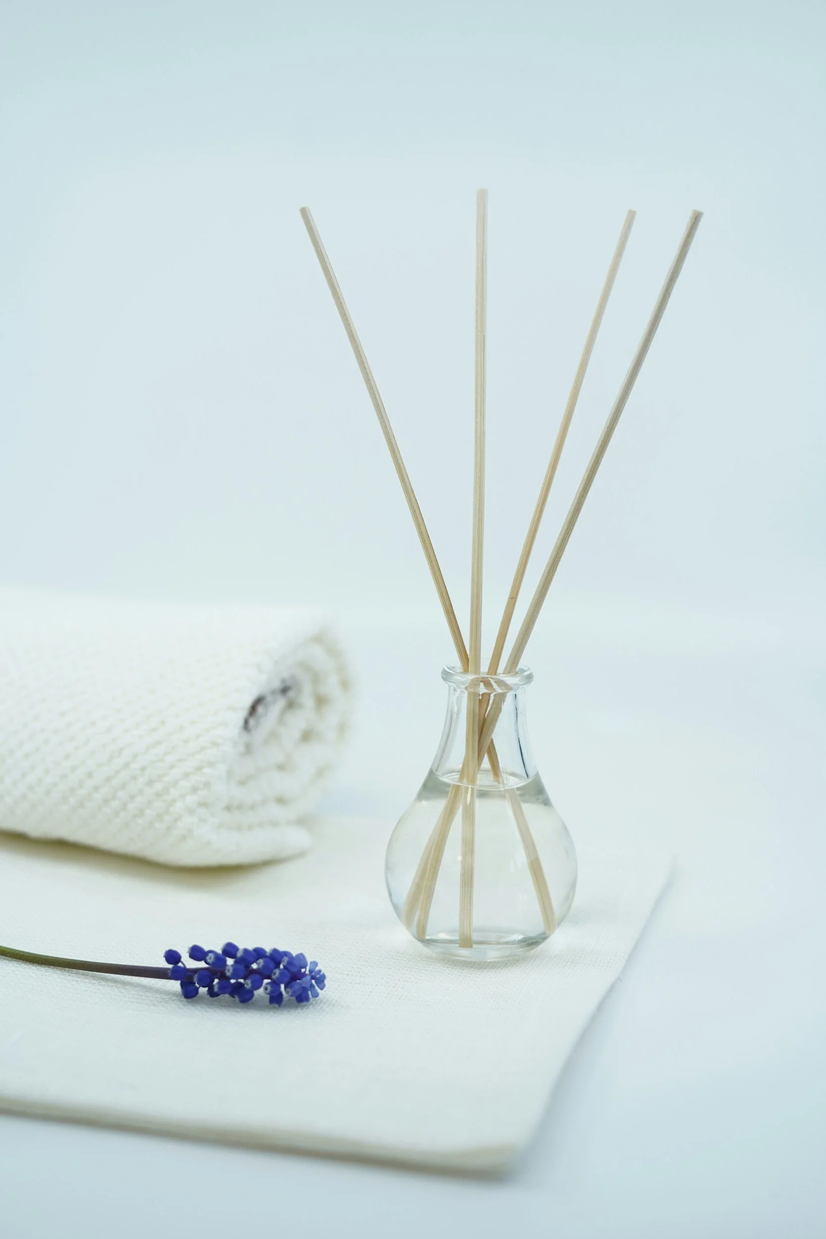 A small glass vase with reed diffusers, a white towel, and a sprig of purple flowers on a white surface.
