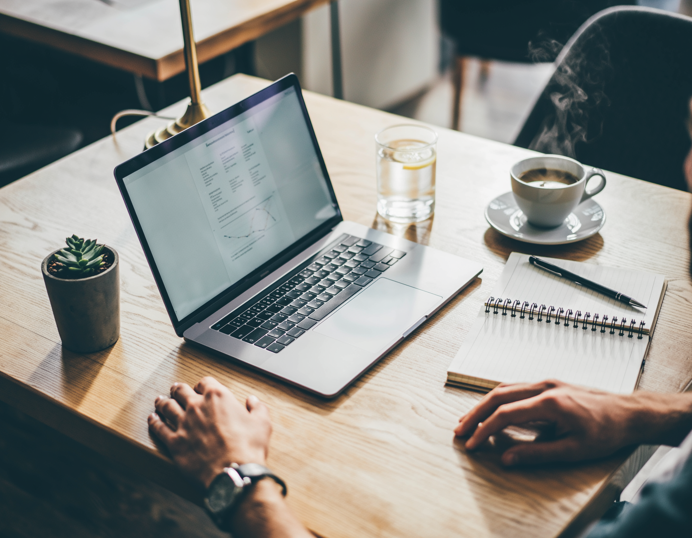 A laptop displaying a graph, a notebook with a pen, a cup of coffee, a glass of water with lemon, and a small potted succulent on a wooden table in a cozy workspace.
