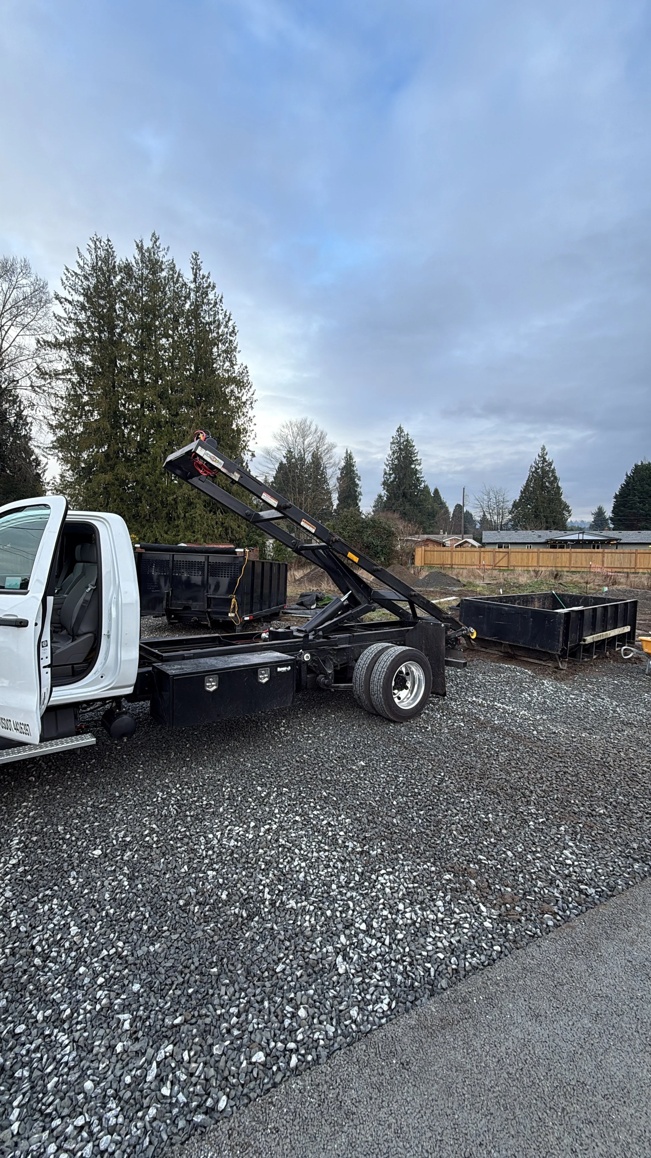 A white flatbed tow truck with a black hydraulic lift parked on a gravel surface, with several empty black drainage or trash bins nearby, outdoors under overcast skies with trees and homes in the background.