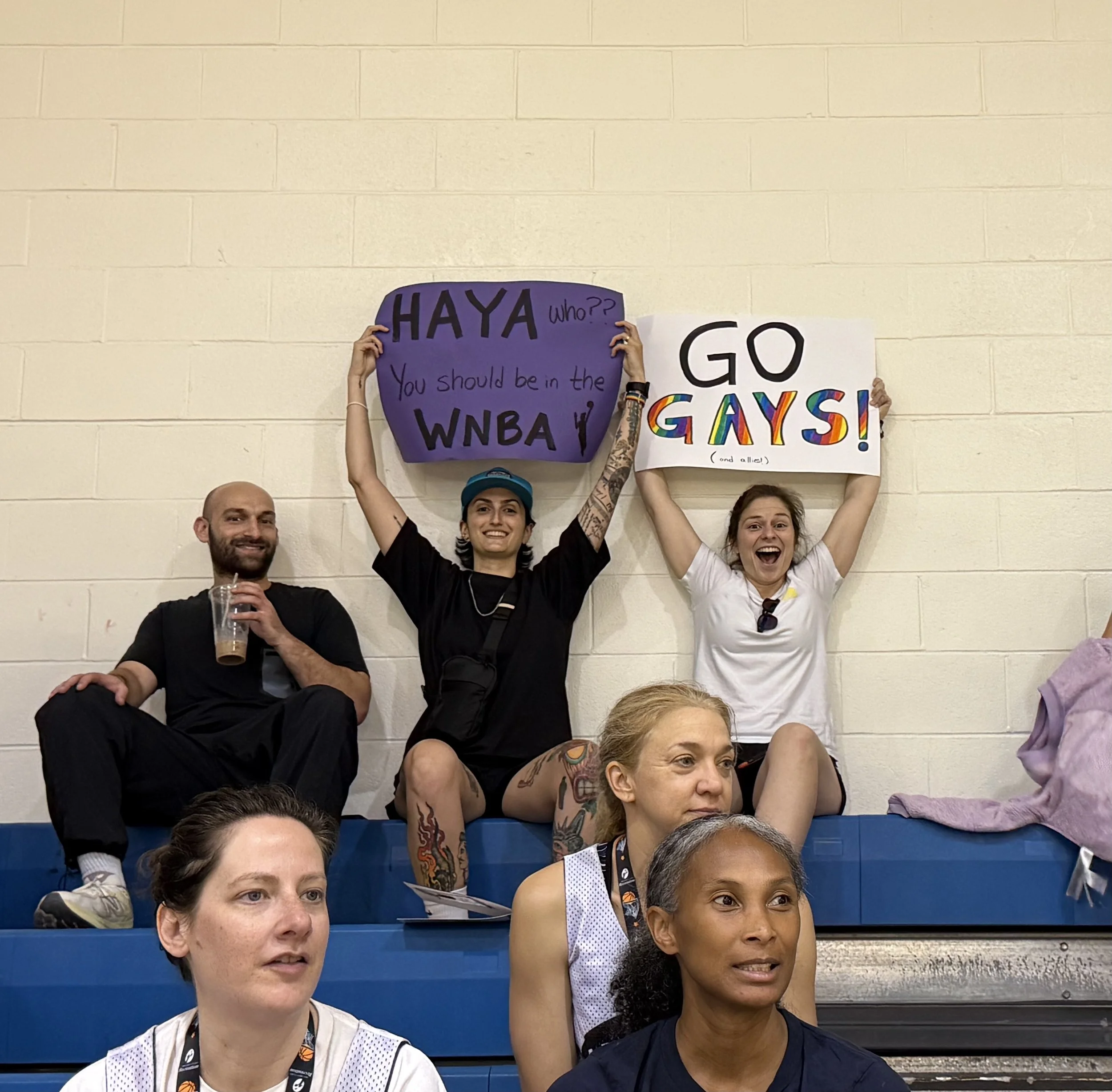 Group of people sitting on bleachers, two people in the back holding up signs, one with rainbow-colored letters that says 'GO GAYS!' and another with purple background and black text supporting WNBA. Others look on with neutral or amused expressions.