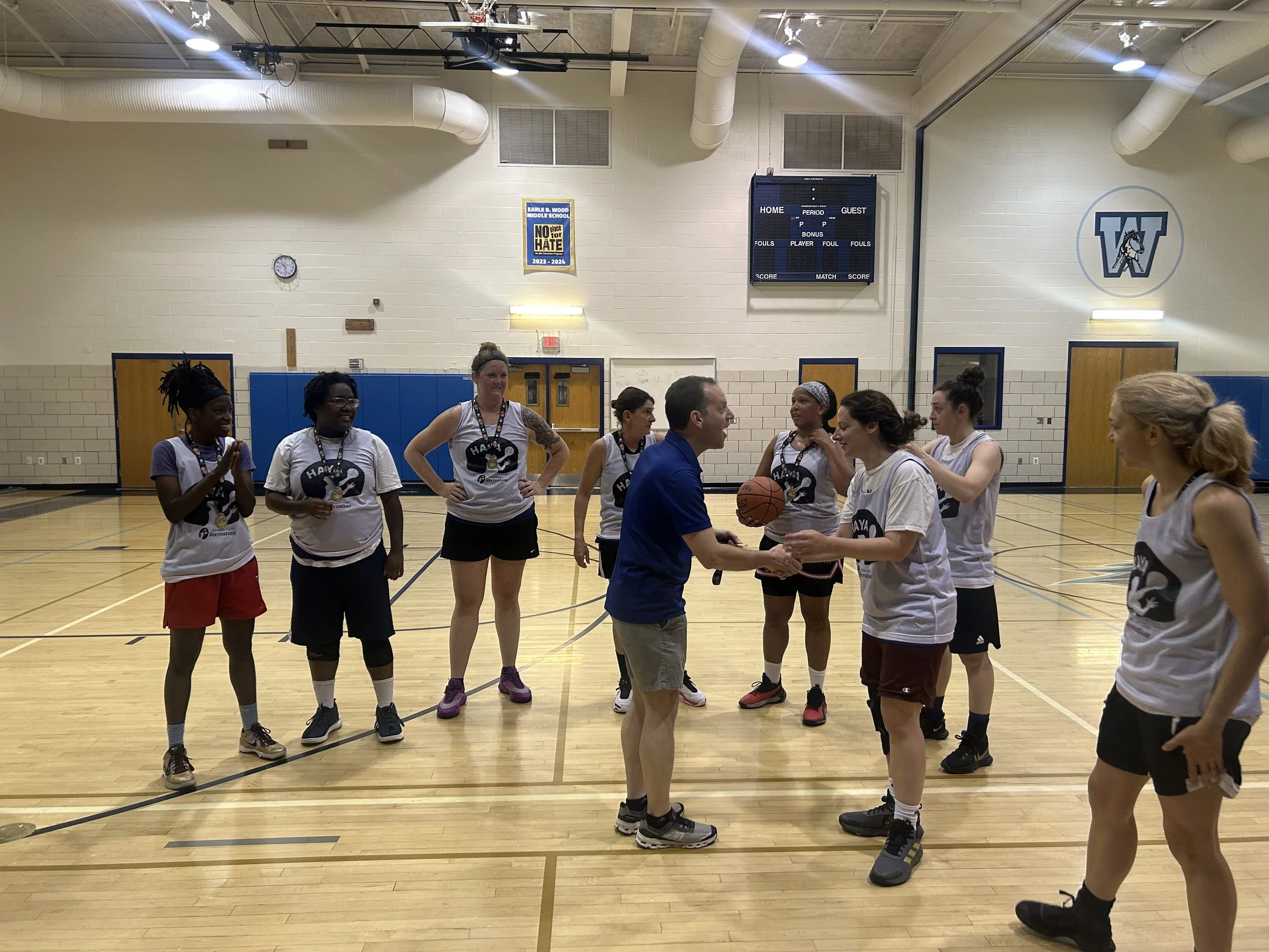 A group of female basketball players in an indoor gym, receiving a trophy from an administrator. They are wearing matching tank tops with a logo, and some are smiling and clapping. The gym has wooden floors and a scoreboard on the wall.