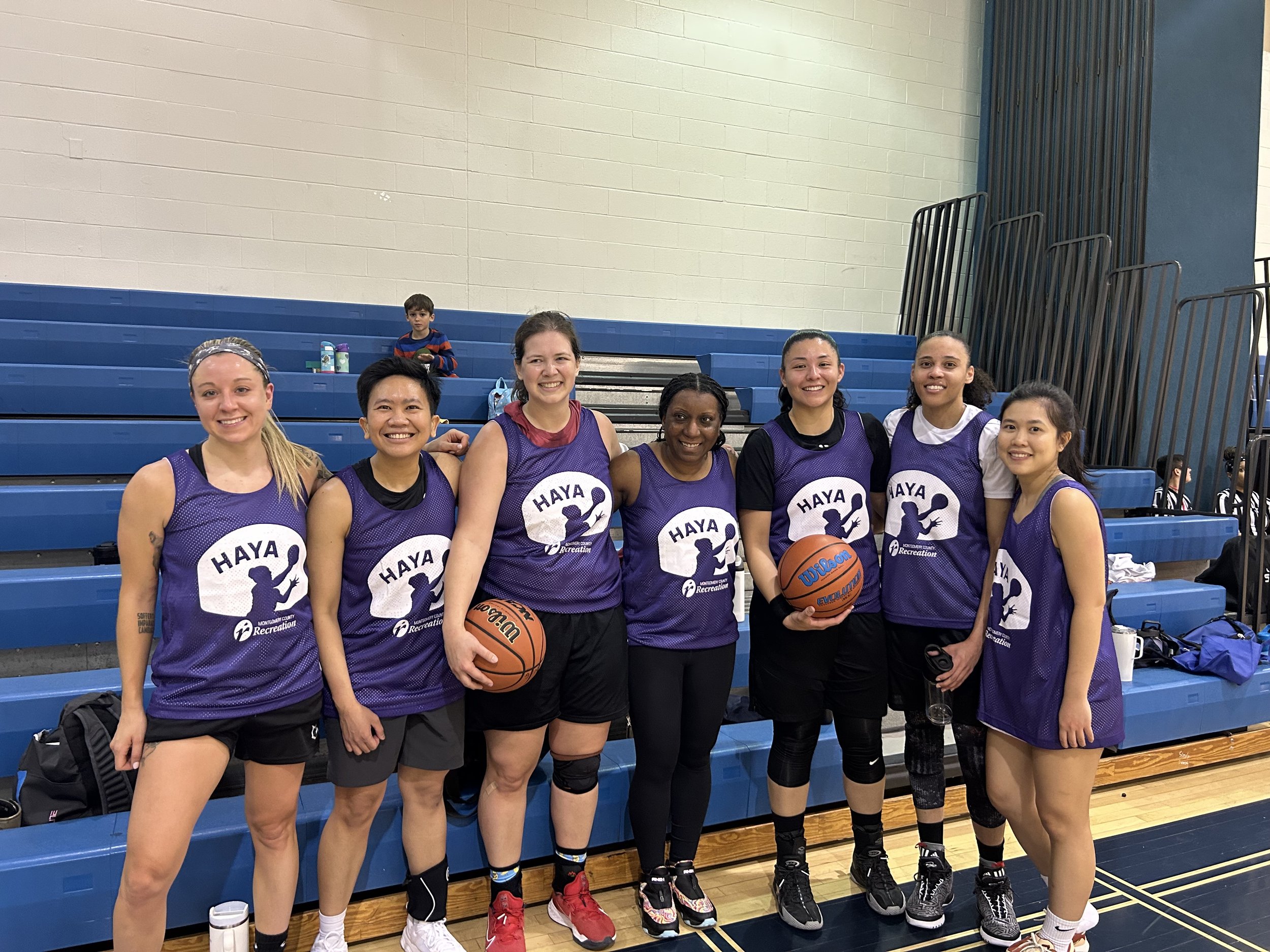 Group of seven women in purple basketball jerseys posing together in a gymnasium, with two women holding basketballs.
