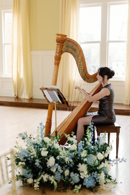 A woman in a black dress playing a harp inside a sunlit room with large windows and yellow curtains, surrounded by a large floral arrangement of white and blue flowers.