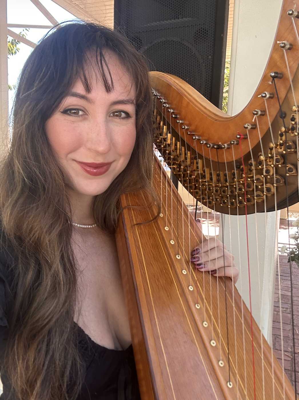 A woman with long brown hair, wearing a black top and pearl necklace, holding a harp.
