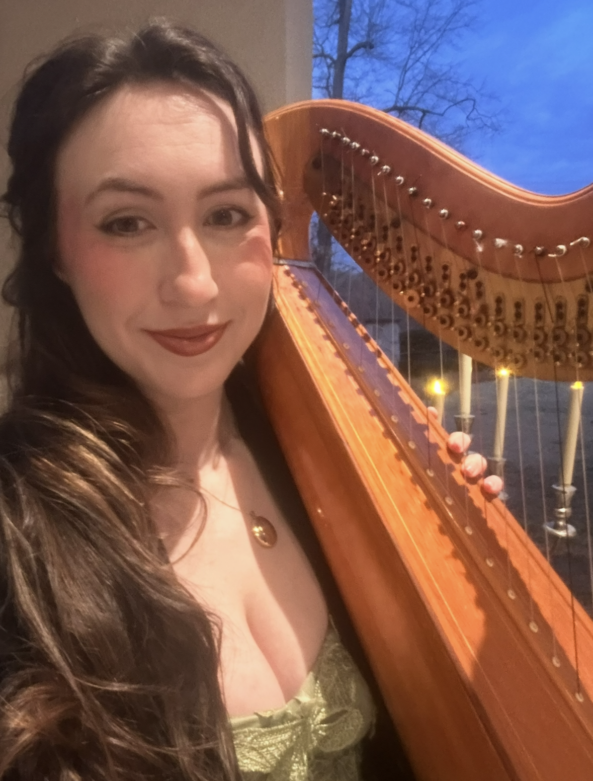 A woman with long brown hair and wearing a gold dress, holding a harp, with candleholders in the background and a blue sky outside.