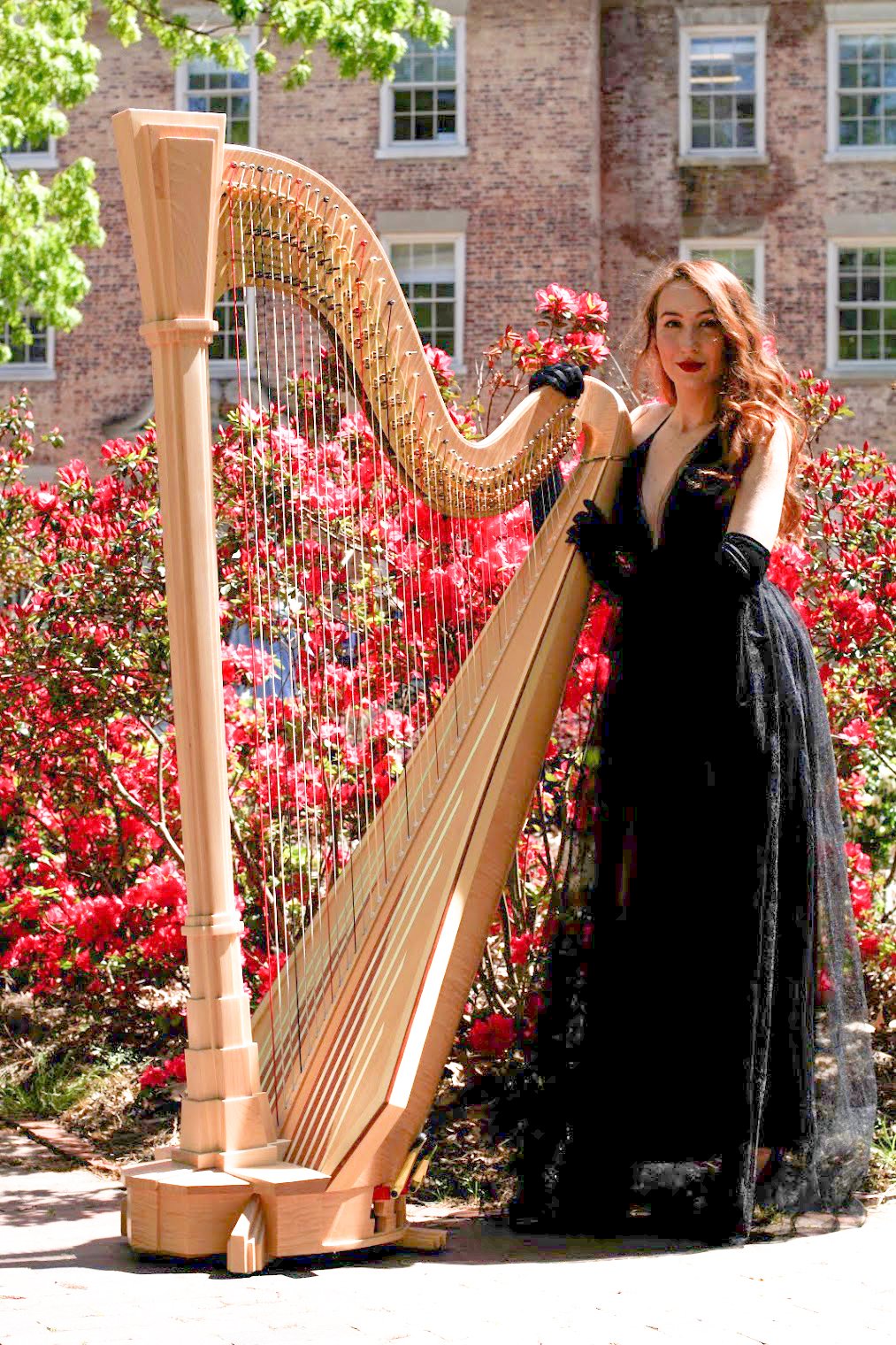 A woman in a black dress and gloves standing next to a harp outdoors, surrounded by pink flowering bushes and a brick building.