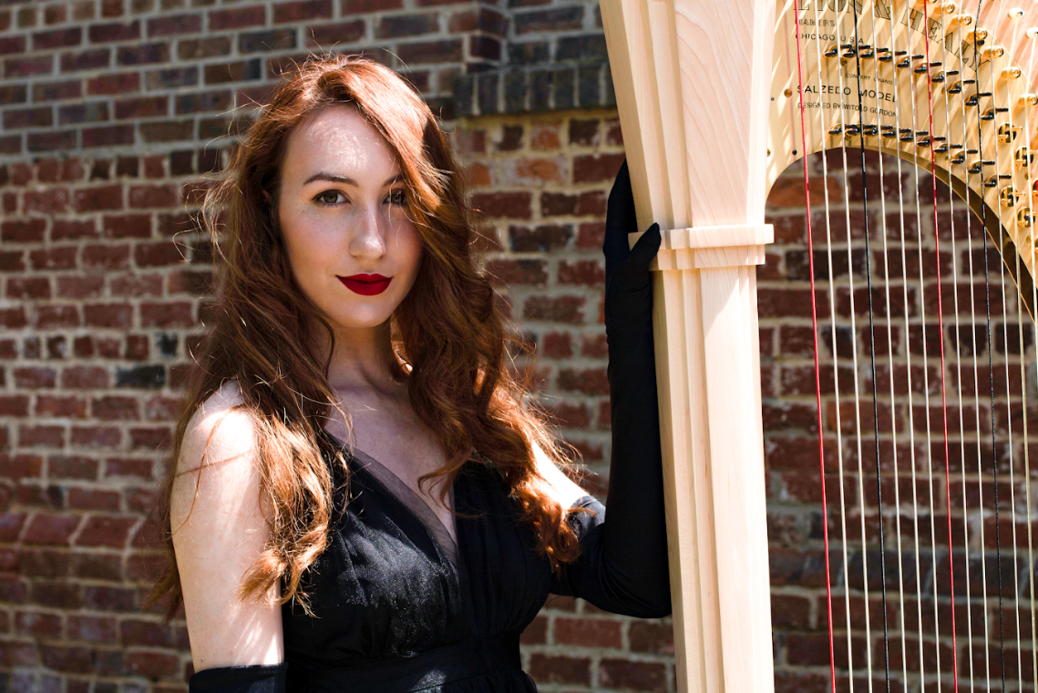 A woman with long, curly red hair and red lipstick standing next to a wooden harp against a brick wall.