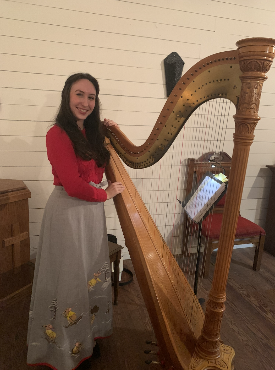 A woman in a red top and a long gray skirt with animal embroidery is standing next to a large wooden harp, smiling at the camera inside a room with white wooden-paneled walls.