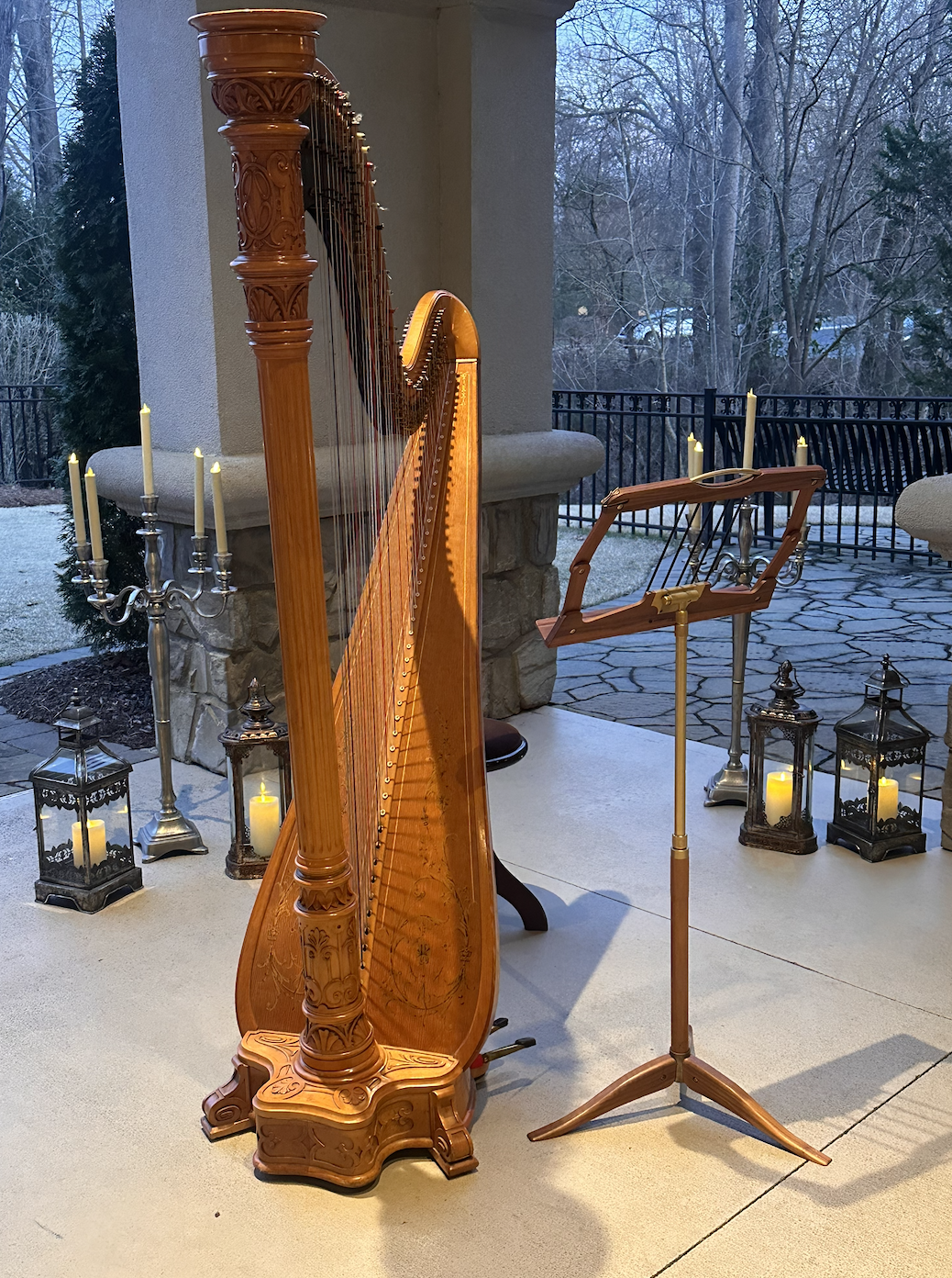 A wooden harp on a porch surrounded by candelabras with lit candles, set against a background of leafless trees and a black metal railing.