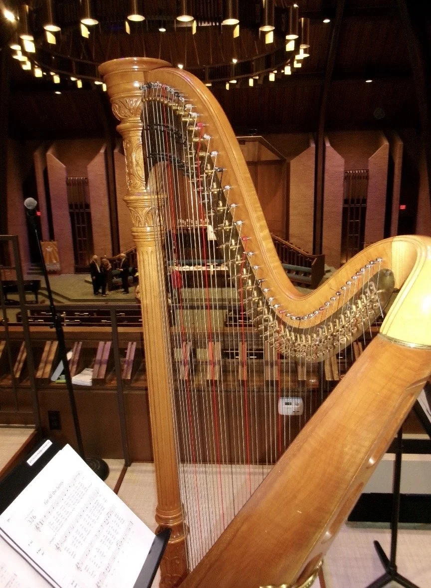 A wooden harp with an open soundboard, set in a concert hall or church setting with people in the background and a music stand with sheet music in the foreground.