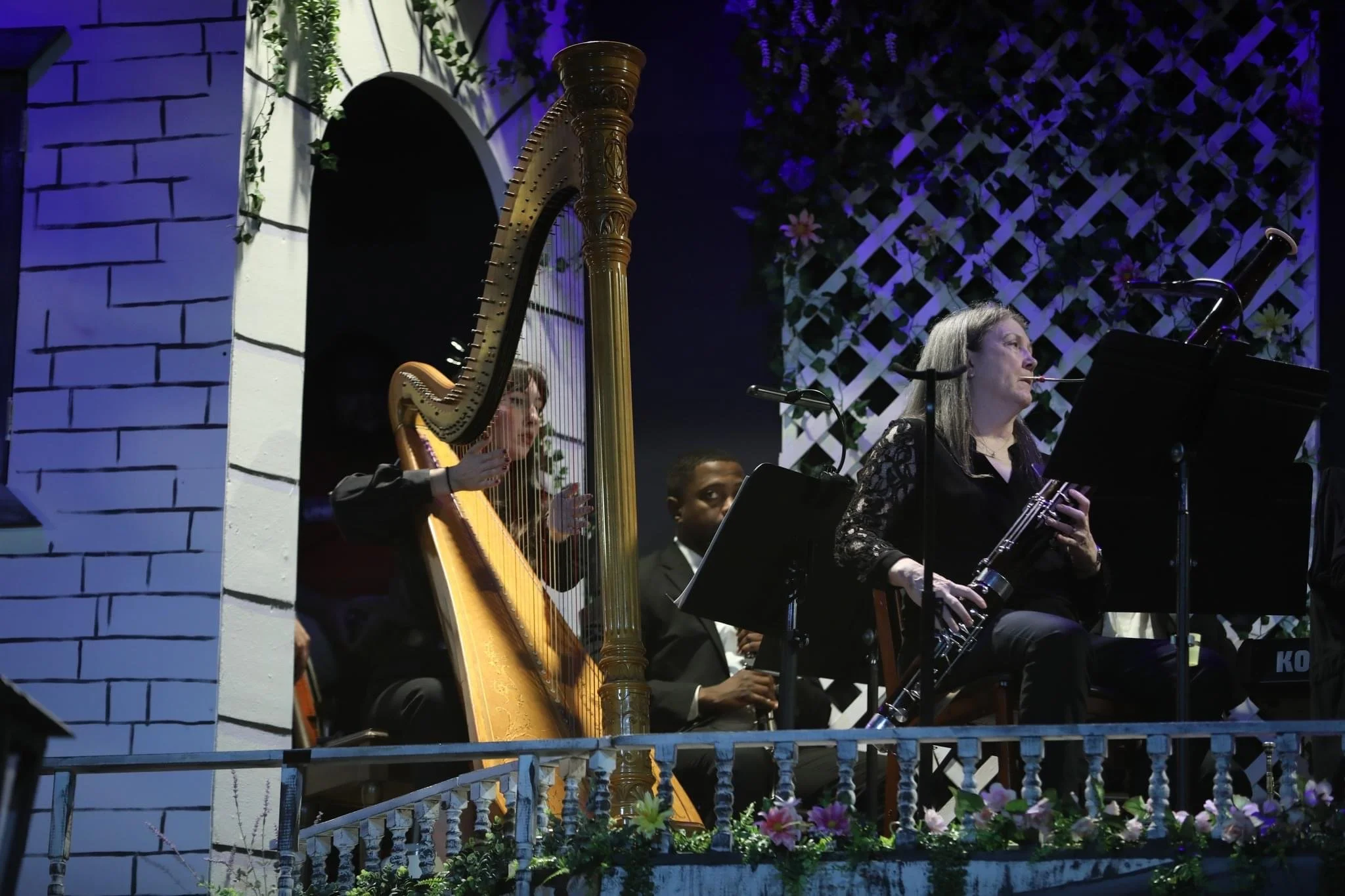 Musicians performing on an outdoor stage with flower decorations, featuring a woman playing the clarinet and a woman playing the harp.