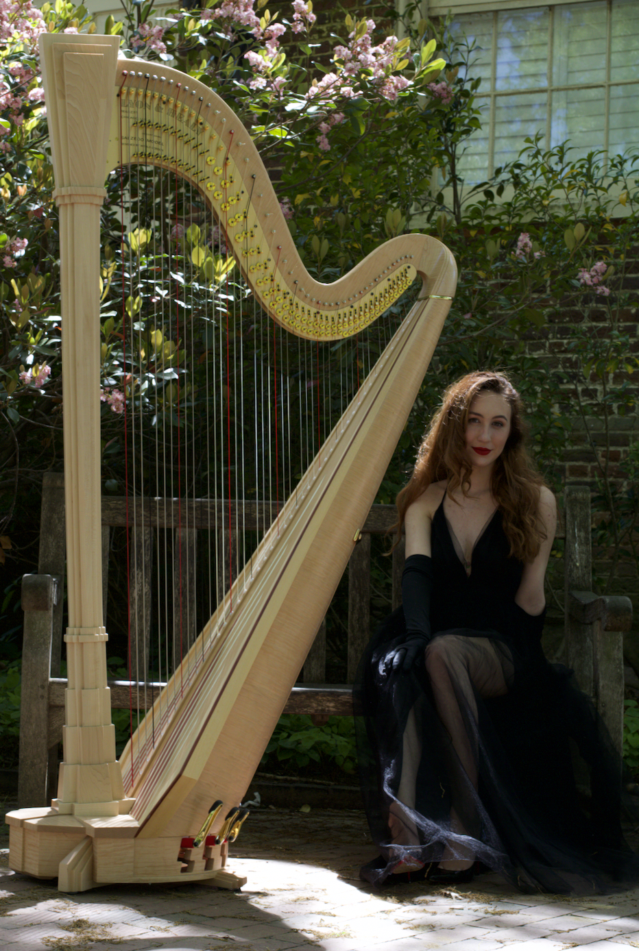A woman with long, curly brown hair and red lipstick sitting on a wooden garden bench next to a large wooden harp, surrounded by green plants and pink flowers.