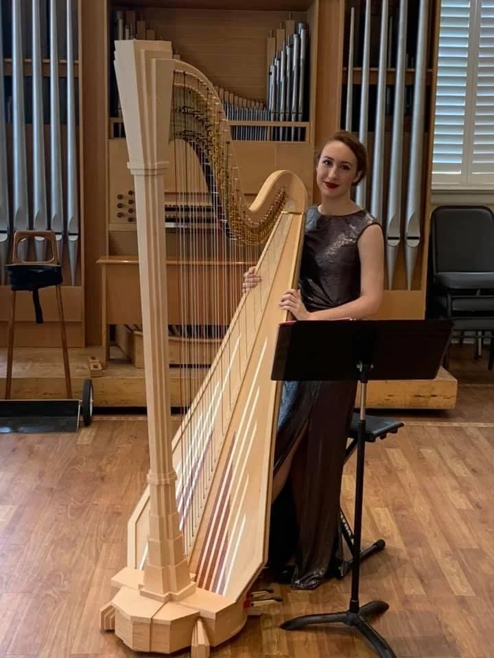 A woman in a black dress standing beside a large wooden harp in a room with wood floors and large organ pipes in the background.