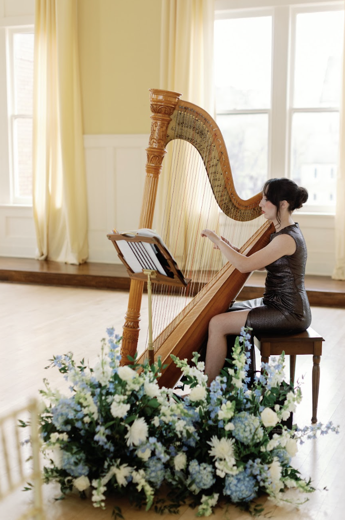 A woman sitting at a harp, reading sheet music, in a bright room with large windows and cream-colored curtains, surrounded by a floral arrangement at her feet.