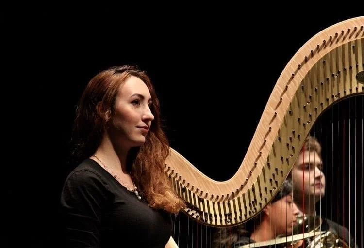 A woman with long brown hair playing a large wooden harp on a dark stage.
