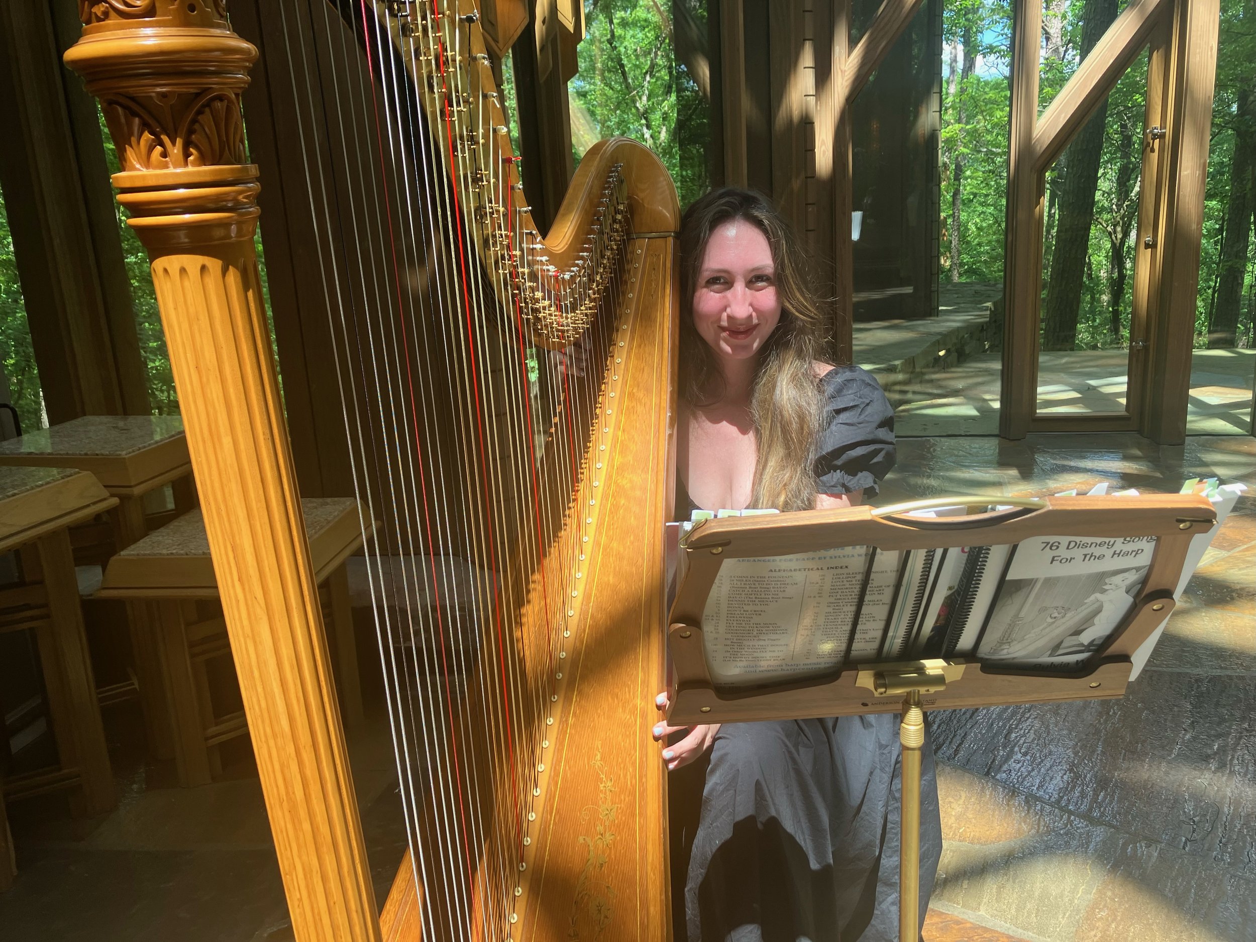 Woman sitting at a music stand next to a harpsichord, inside a wooden structure with glass doors, surrounded by green trees.
