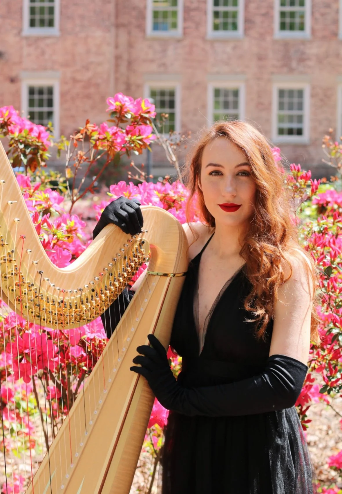 A woman with long red hair and red lipstick in a black dress with long black gloves, posing outdoors with a harp, surrounded by pink azalea flowers.