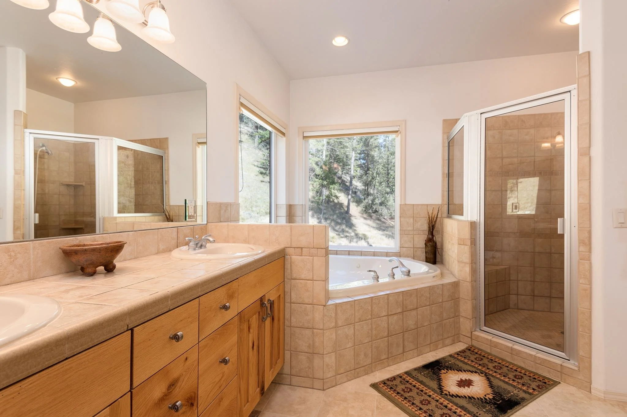 A spacious bathroom with dual sinks, a large mirror, a soaking tub next to a window, and a separate glass-enclosed shower with beige tile walls.