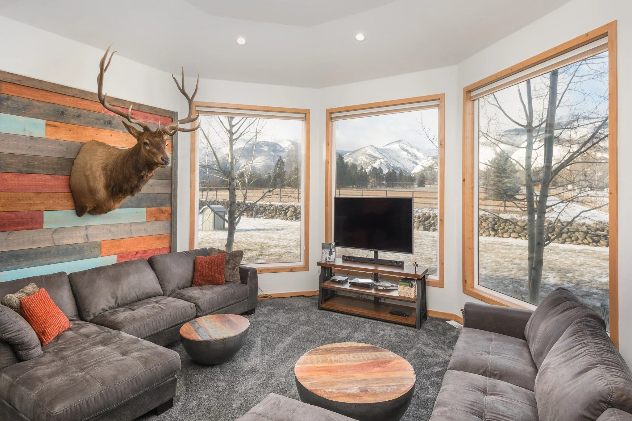 Living room with large windows showing snowy mountains outside, a mounted deer head on a colorful wooden wall, a gray sectional sofa with orange pillows, and a TV on a stand.