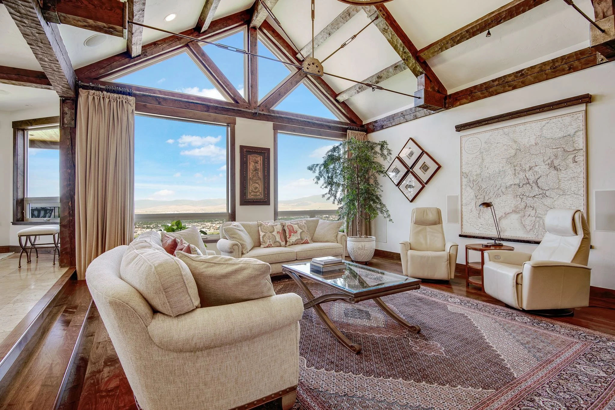 Living room with large windows, wooden beams, cream-colored sofas, white leather armchairs, a coffee table, a large map on the wall, and a potted plant.