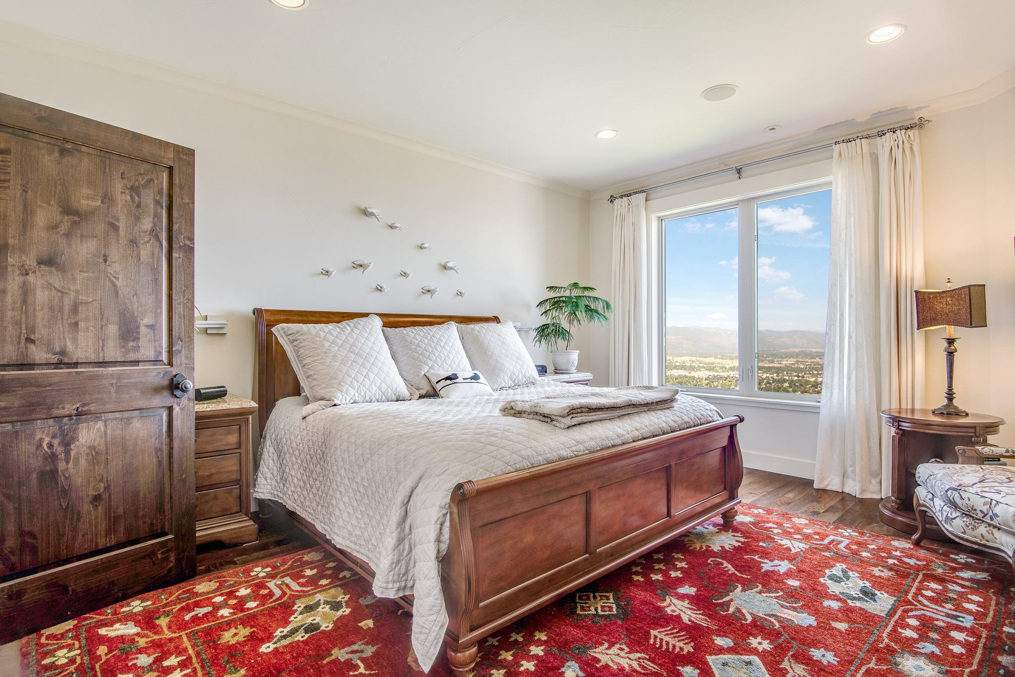 Bedroom with wooden bed, white bedding, pillows, a large window showing landscape, a potted plant, a nightstand, lamp, and a red patterned rug.