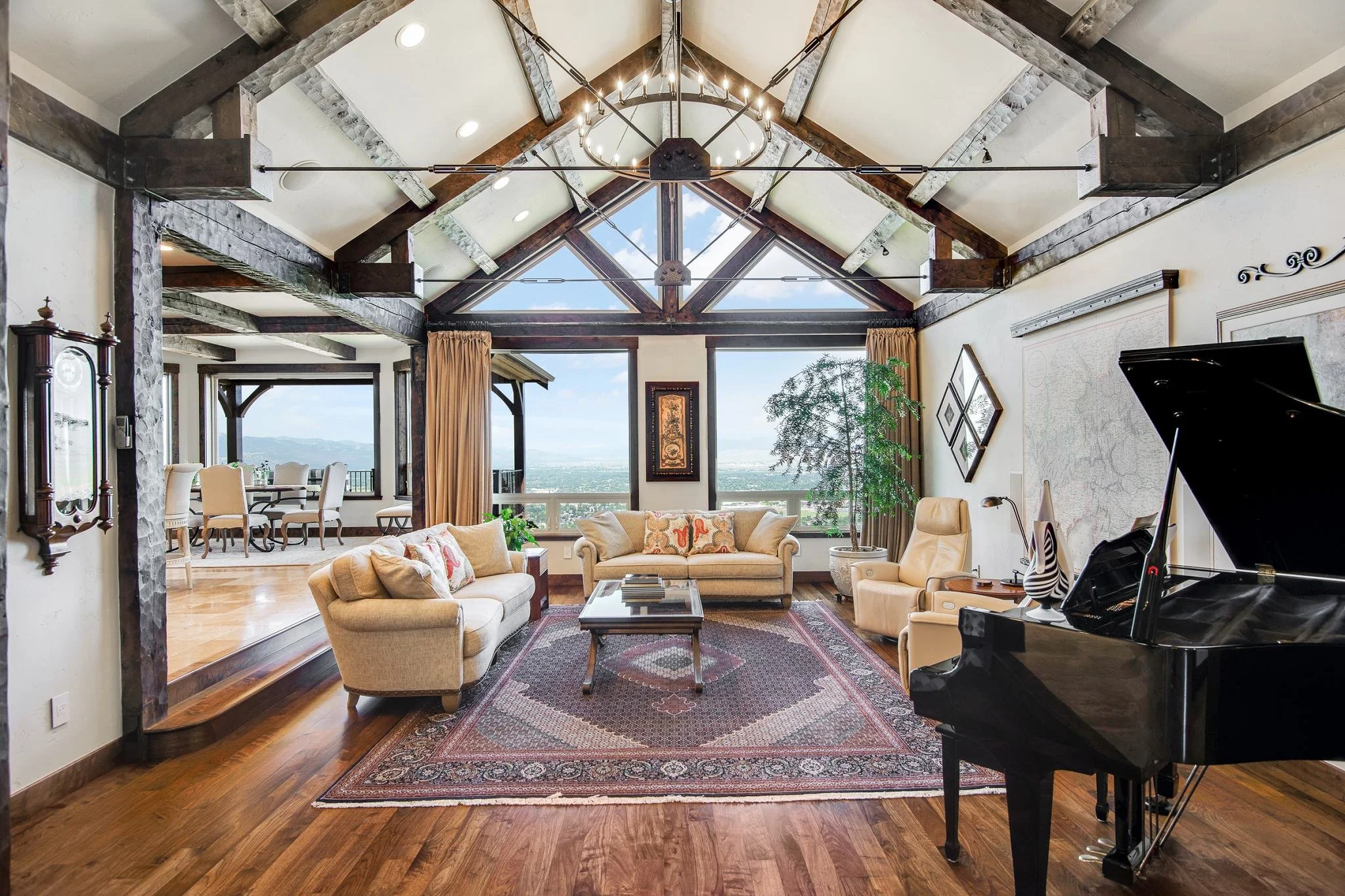 Living room with high vaulted ceiling with exposed wood beams, large window with curtains, beige sofas, a grand piano, and an area rug.