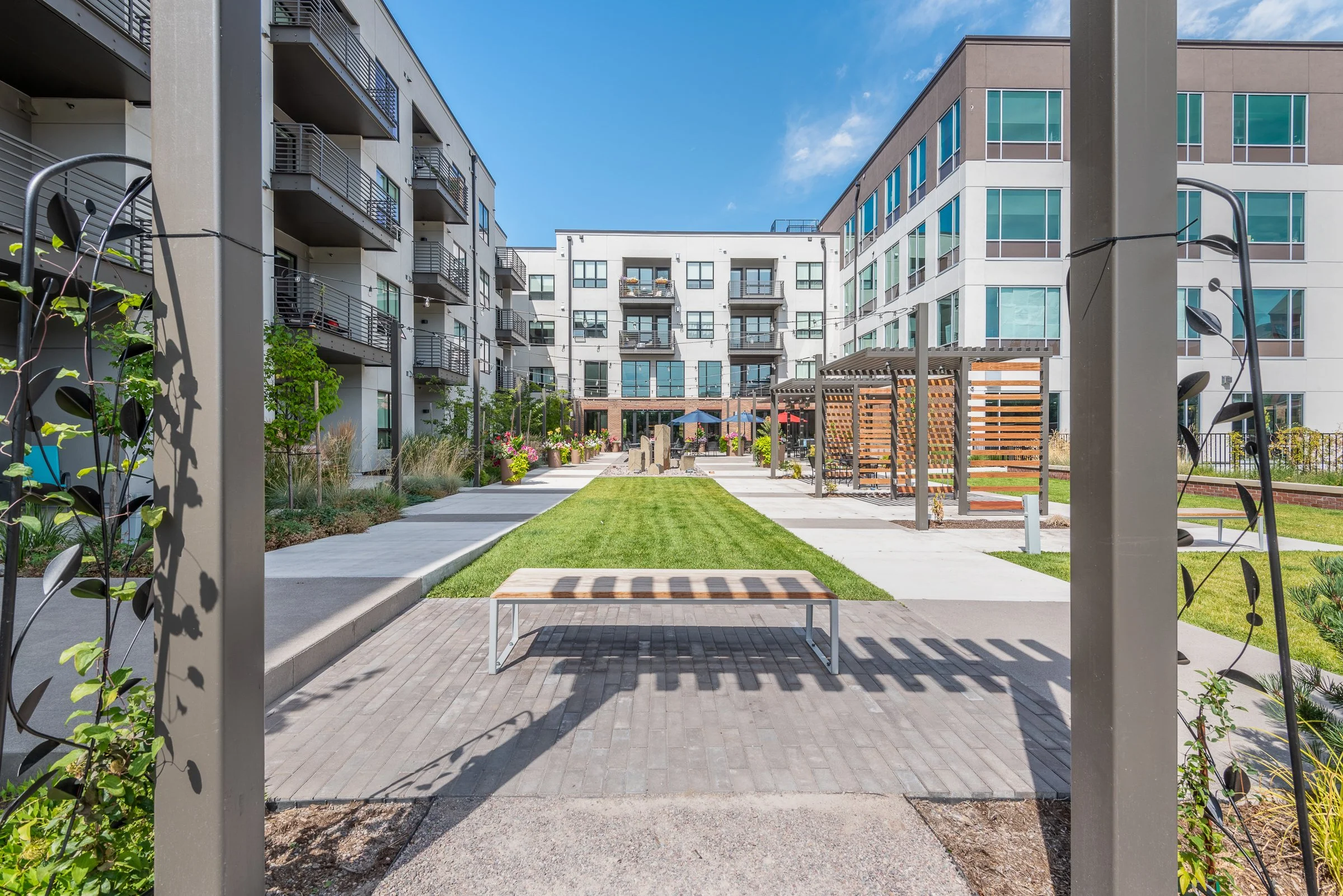 Courtyard of a modern apartment complex with a central grassy area, concrete walkways, benches, plants, and outdoor seating under umbrellas, surrounded by multi-story buildings.