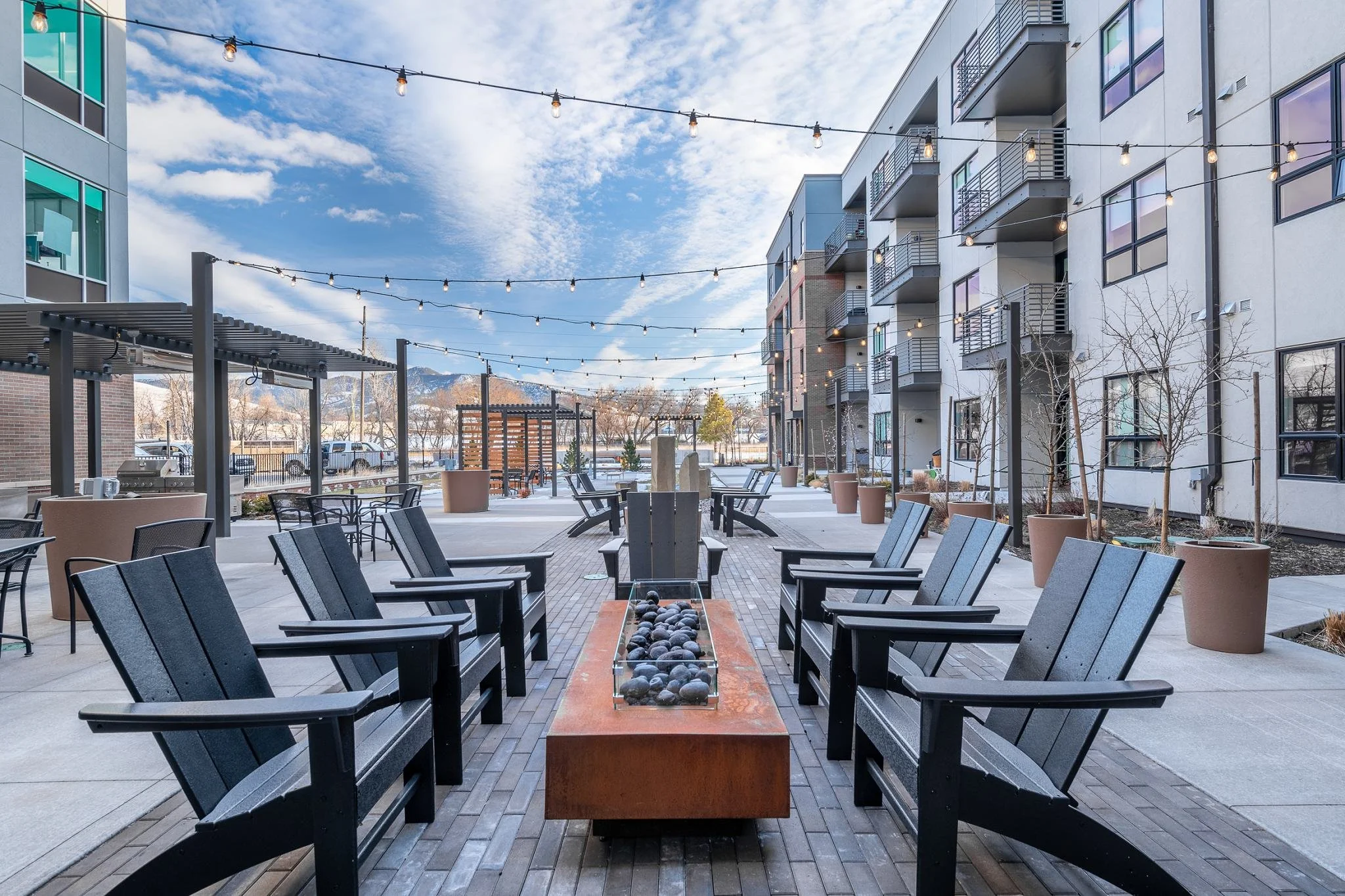 Outdoor lounge area with black chairs around a rectangular fire pit, string lights overhead, modern apartment buildings on the right, and potted trees along the walkway, under a partly cloudy sky.