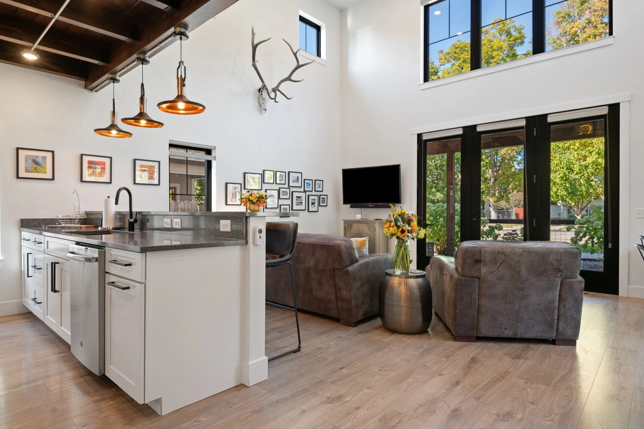 Open living room with high ceiling, large windows showing trees outside, a TV on a stand, brown sofas, armchairs, a kitchen island with black hardware, hanging lights, wall art, and a deer antler mounted on the wall.