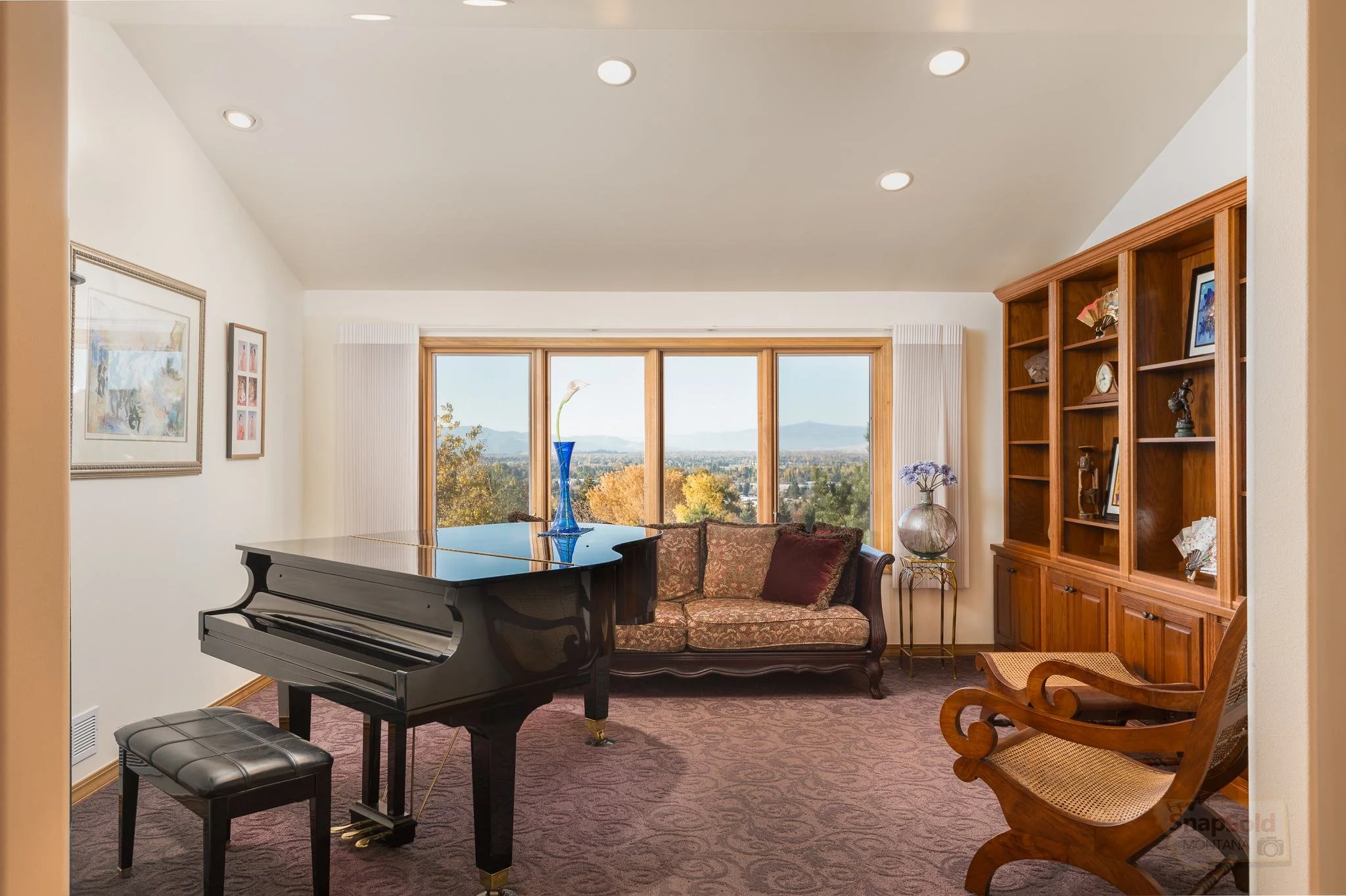 Living room with large window view, upright piano, floral sofa, wooden bookshelf, and decorative items.