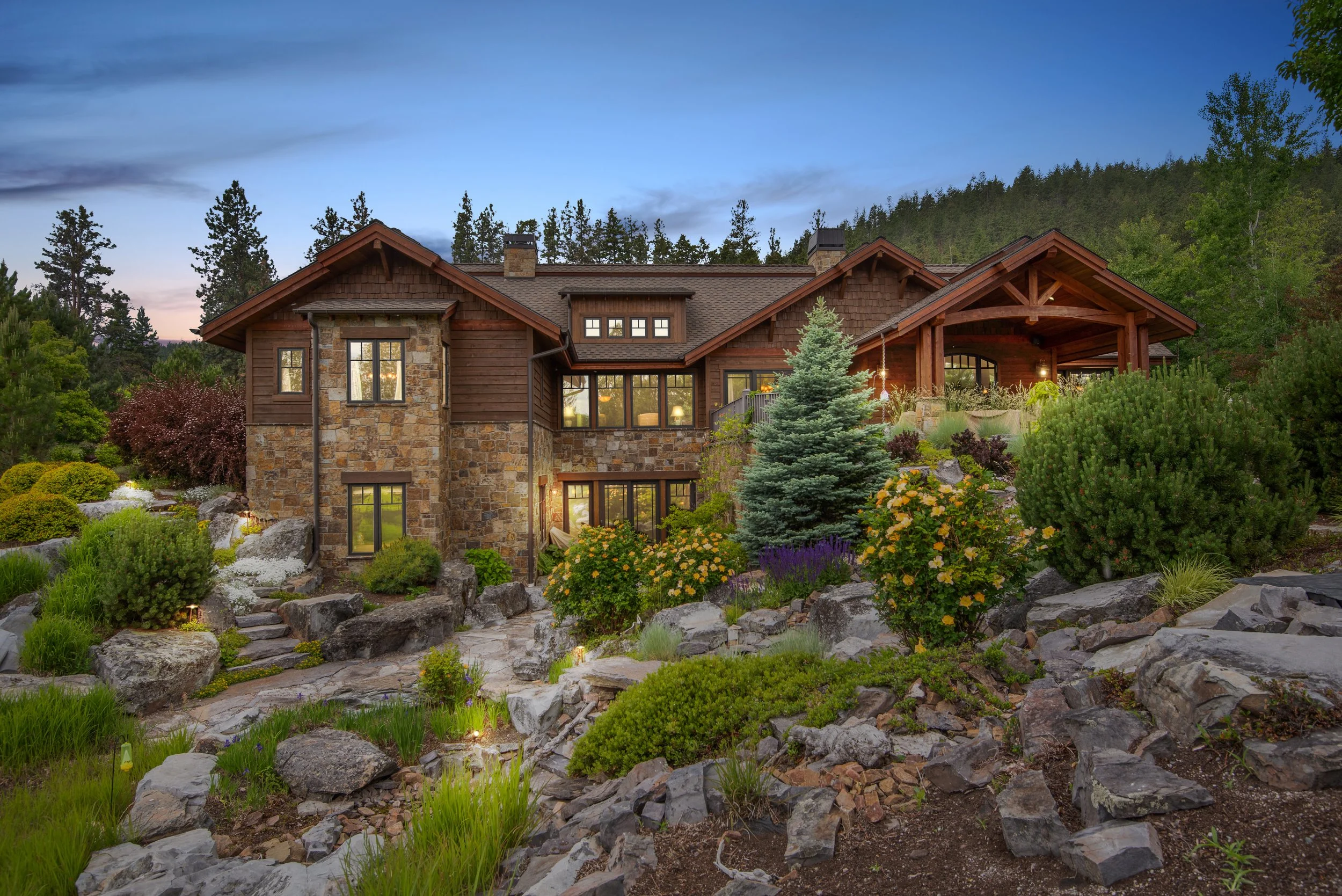 A large house with a stone and wood exterior surrounded by a landscaped garden with rocks, shrubs, and trees, with a backdrop of a forested hillside under a sunset sky.