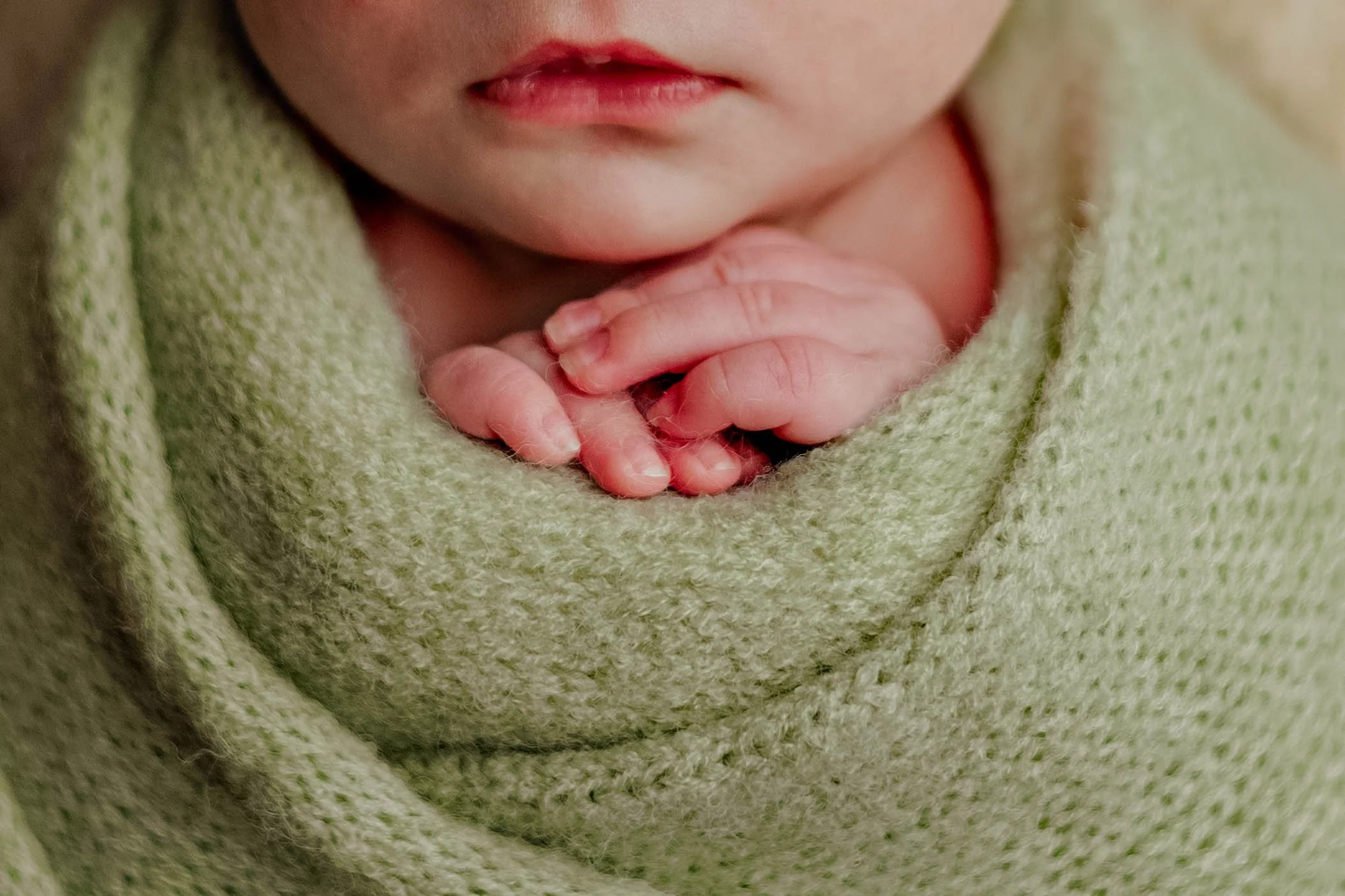 Close-up of a baby's face, showing lips and small hand, wrapped in a green knitted blanket/Wrap.