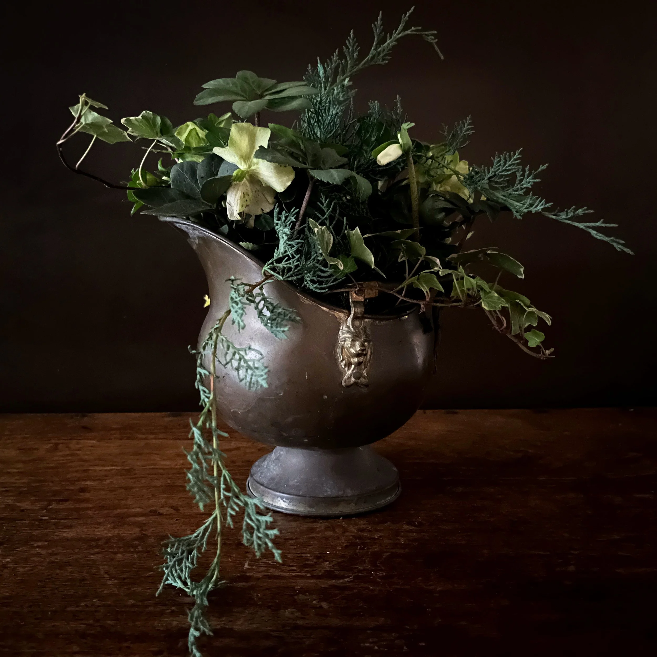 A silver antique vase filled with various green foliage and plants, placed on a wooden surface against a dark background.