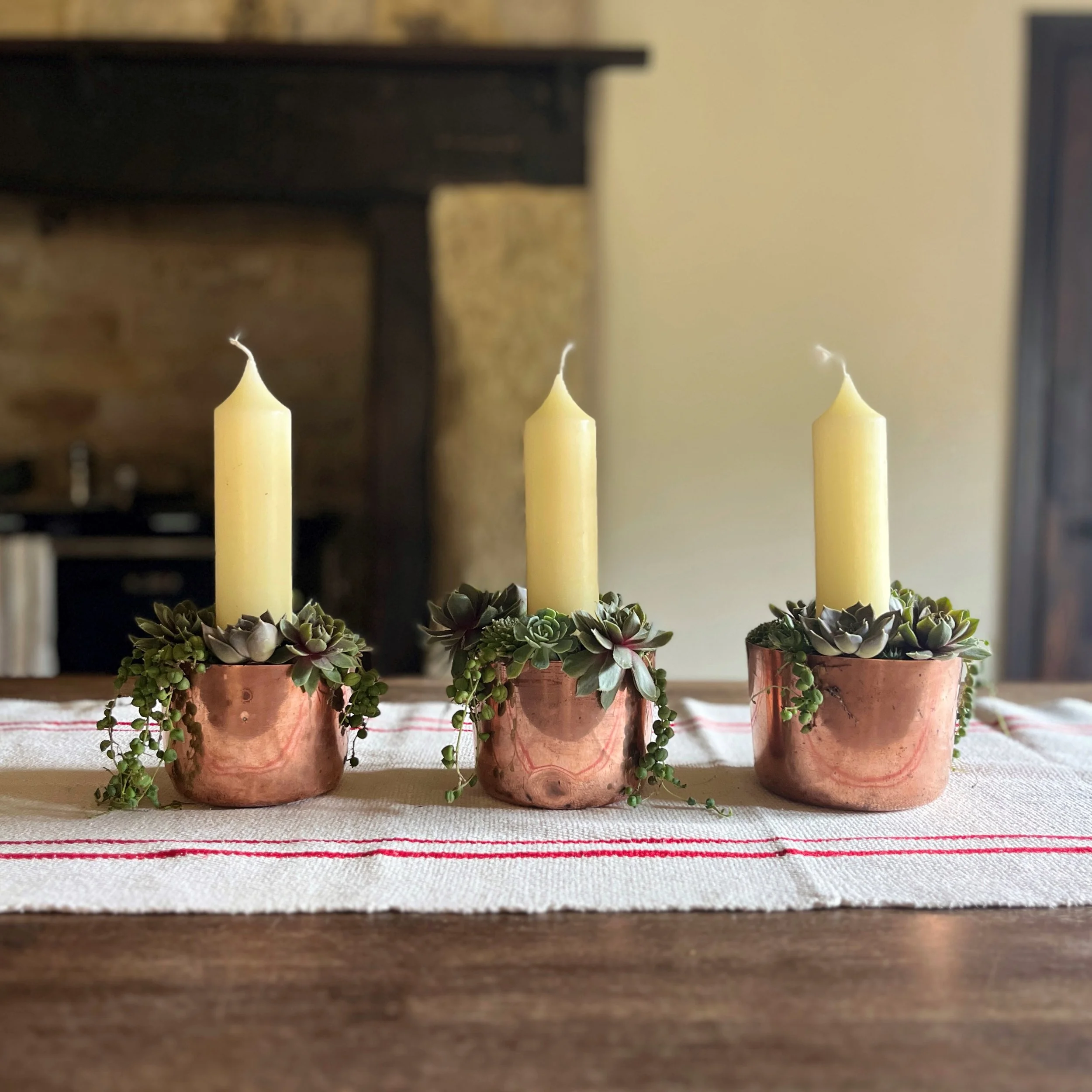 Three copper candle holders with white candles and succulent plants, arranged on a table with a white cloth featuring red stripes, in front of a fireplace.