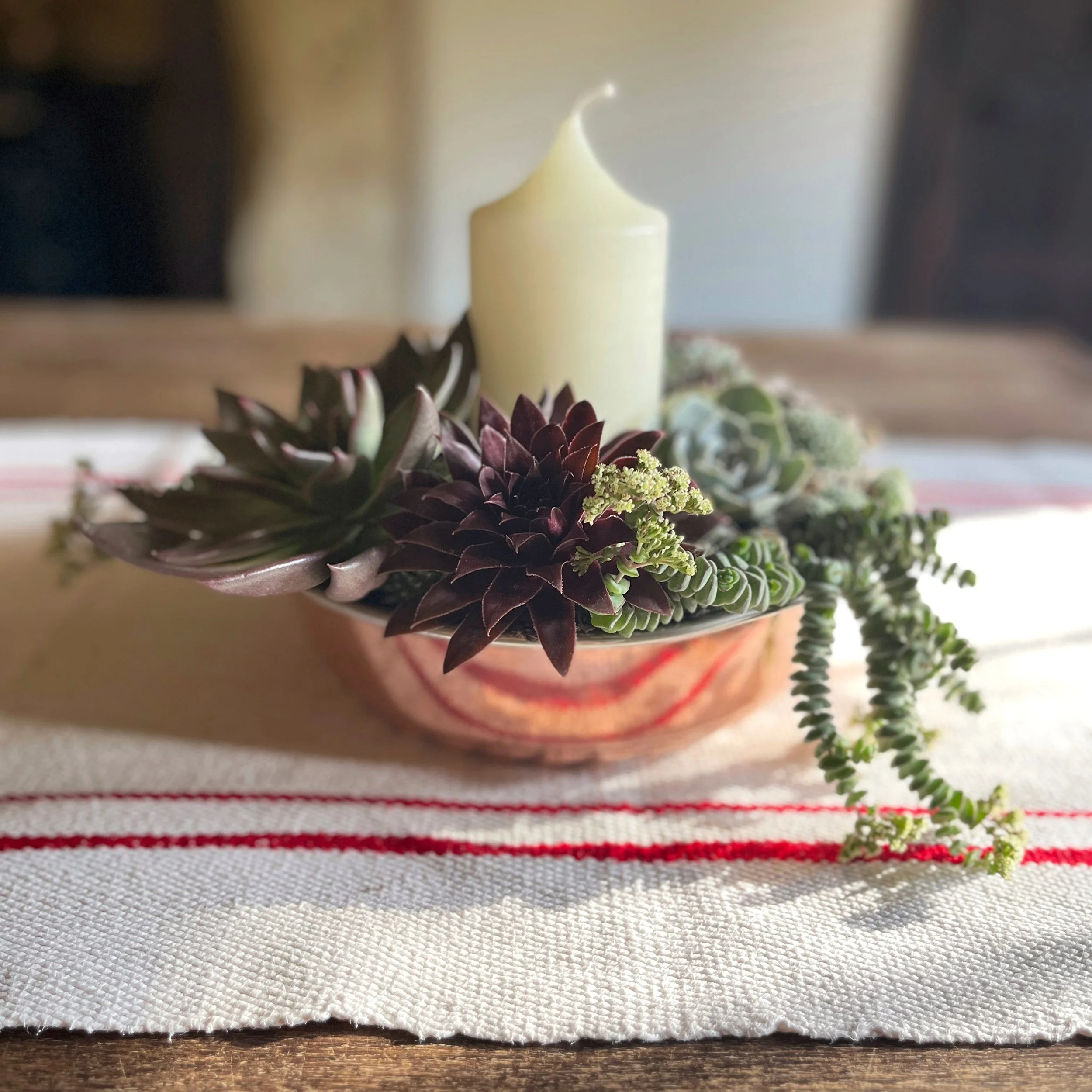 A decorative arrangement of succulents and a white candle in a copper bowl on a woven table runner.