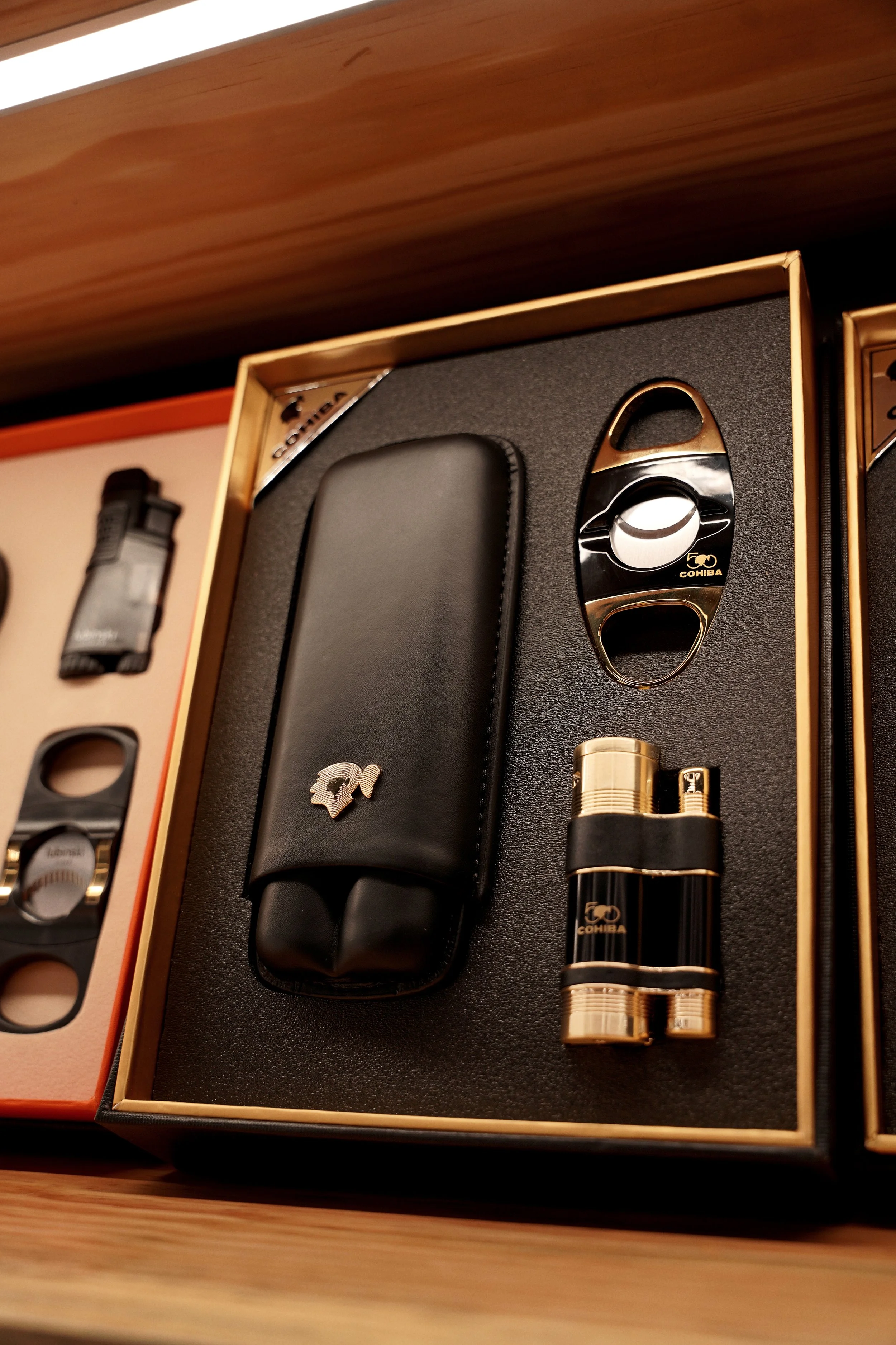 A gift set containing a black leather item with a silver logo, a black and gold disposable lighter, a black and gold cigar cutter, cigar poker