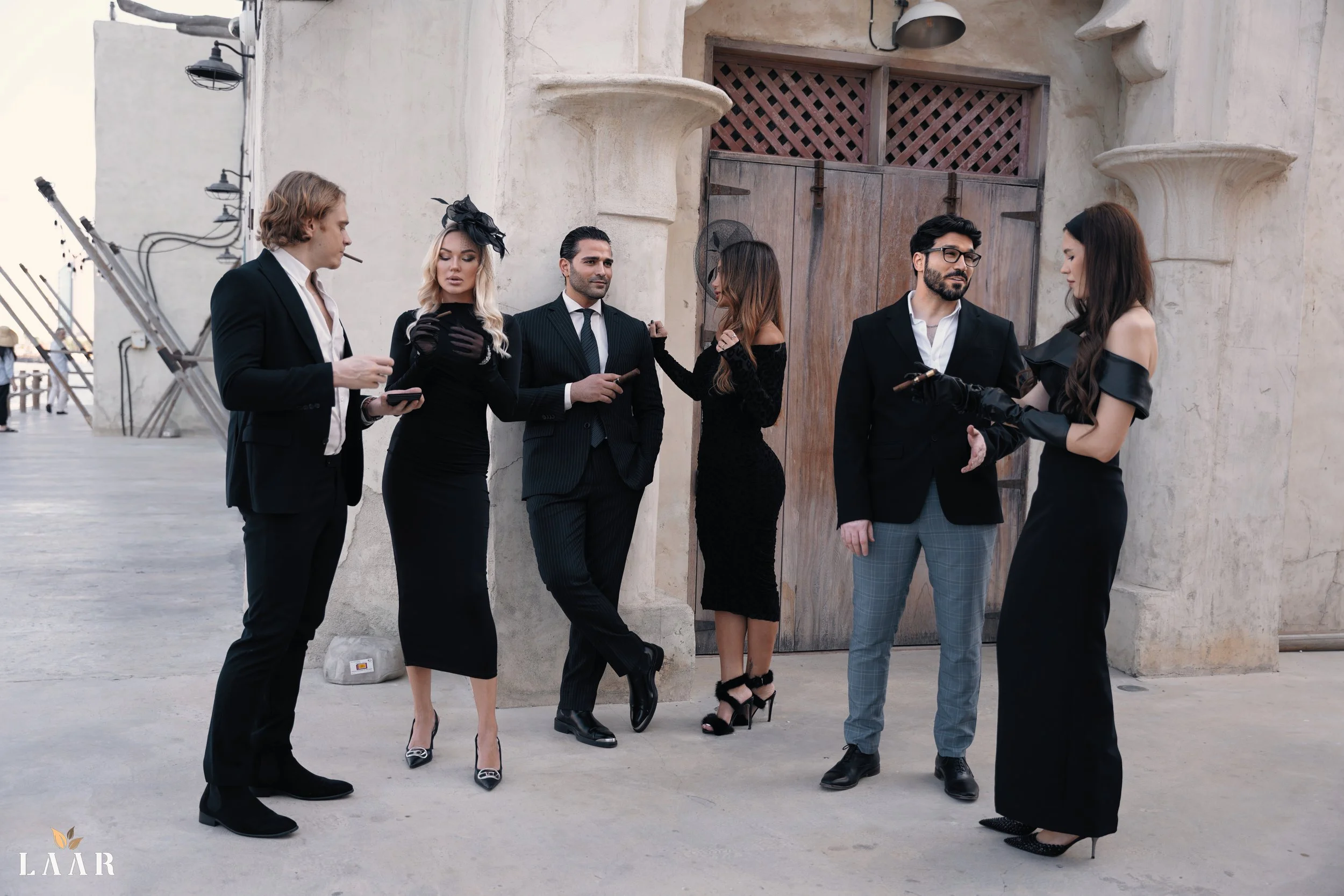 Group of six stylishly dressed people standing outside in front of a rustic door, engaging in conversation and using their phones.
