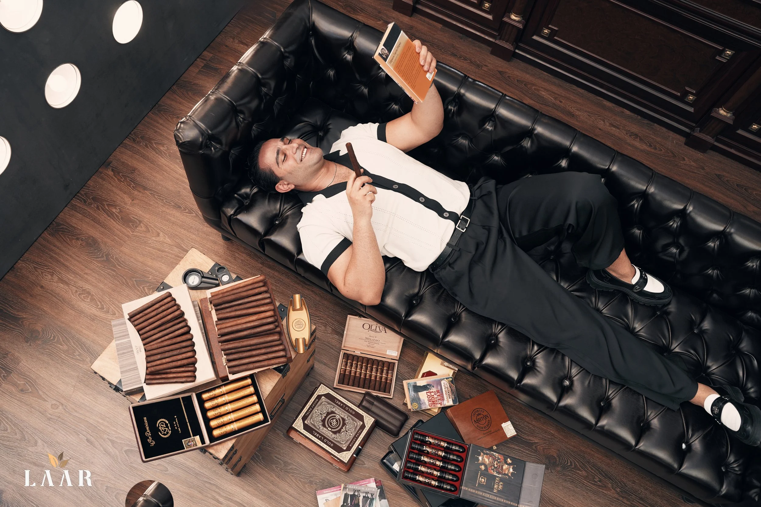 A man lying on a black leather couch surrounded by cigar boxes and cigars, reading a book and holding a cigar in his right hand.