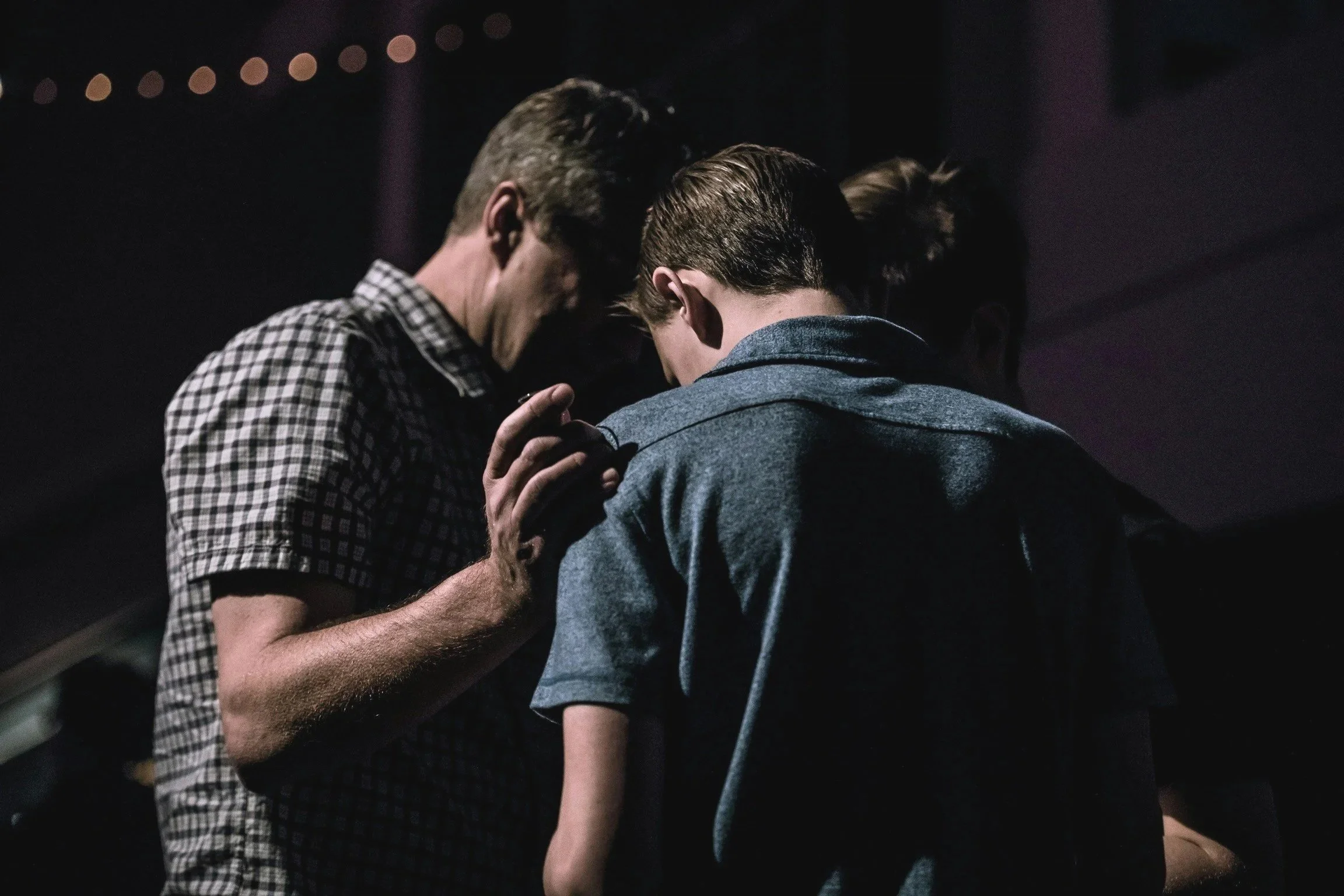 Group of people praying in a dark room with string lights in the background.