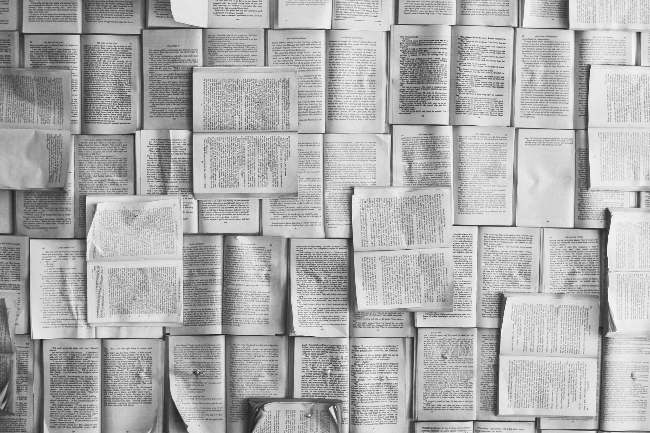A black and white overhead shot of numerous open books with varying sizes and page arrangements, laid out closely together, some overlapping.