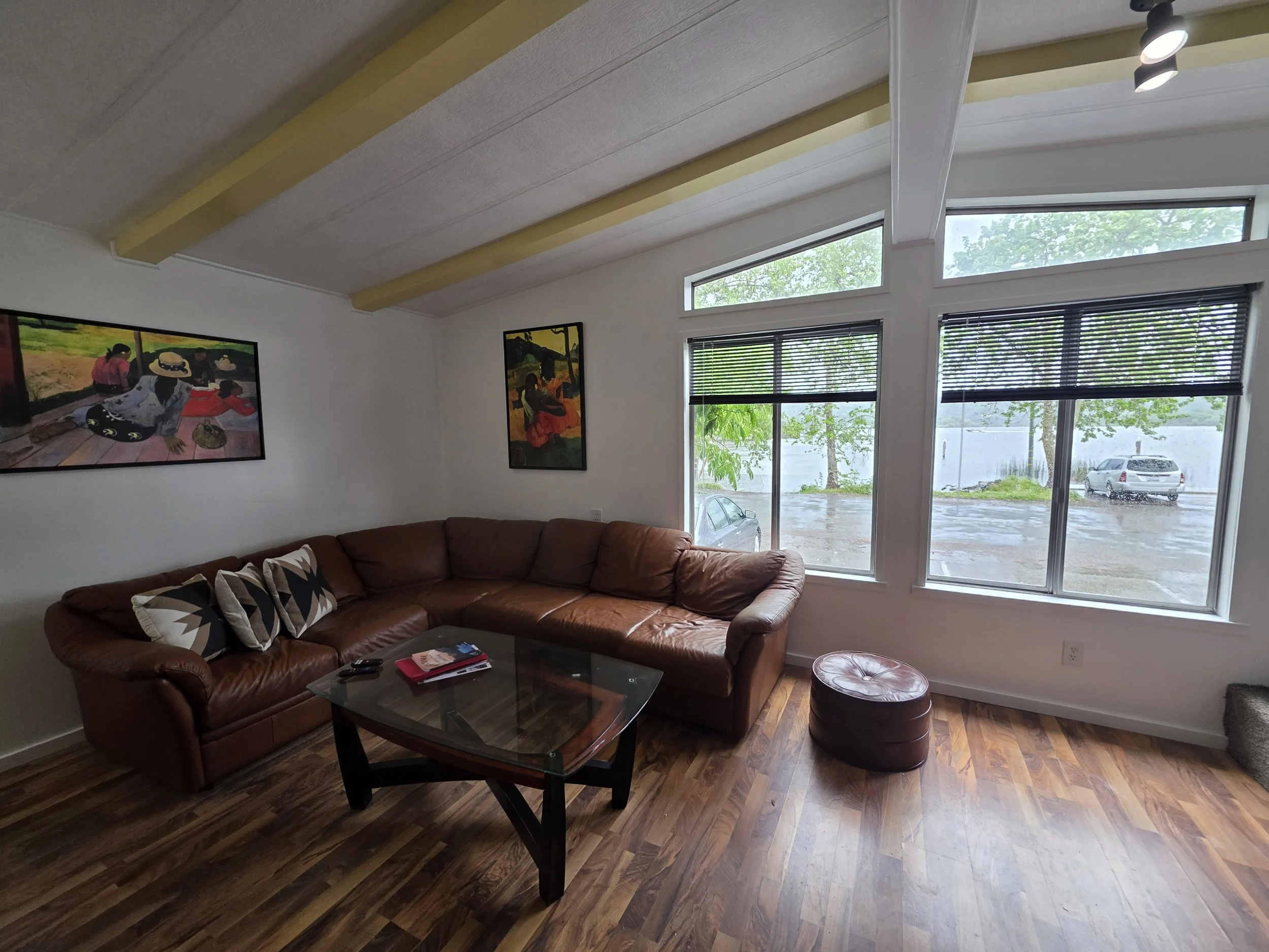 A living room with a large brown leather sectional sofa, decorative pillows, a glass coffee table with magazines, a small leather ottoman, artwork on the white walls, and large windows with blinds showing a rainy day outside with parked cars and tree