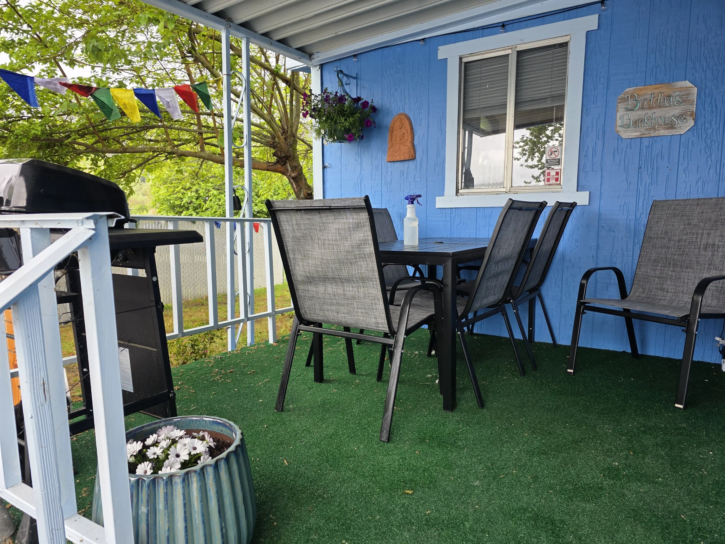 Covered outdoor patio with a black dining table and six chairs, a propane grill, potted flowers, a blue house wall with a window, a wooden sign, and colorful bunting flags.