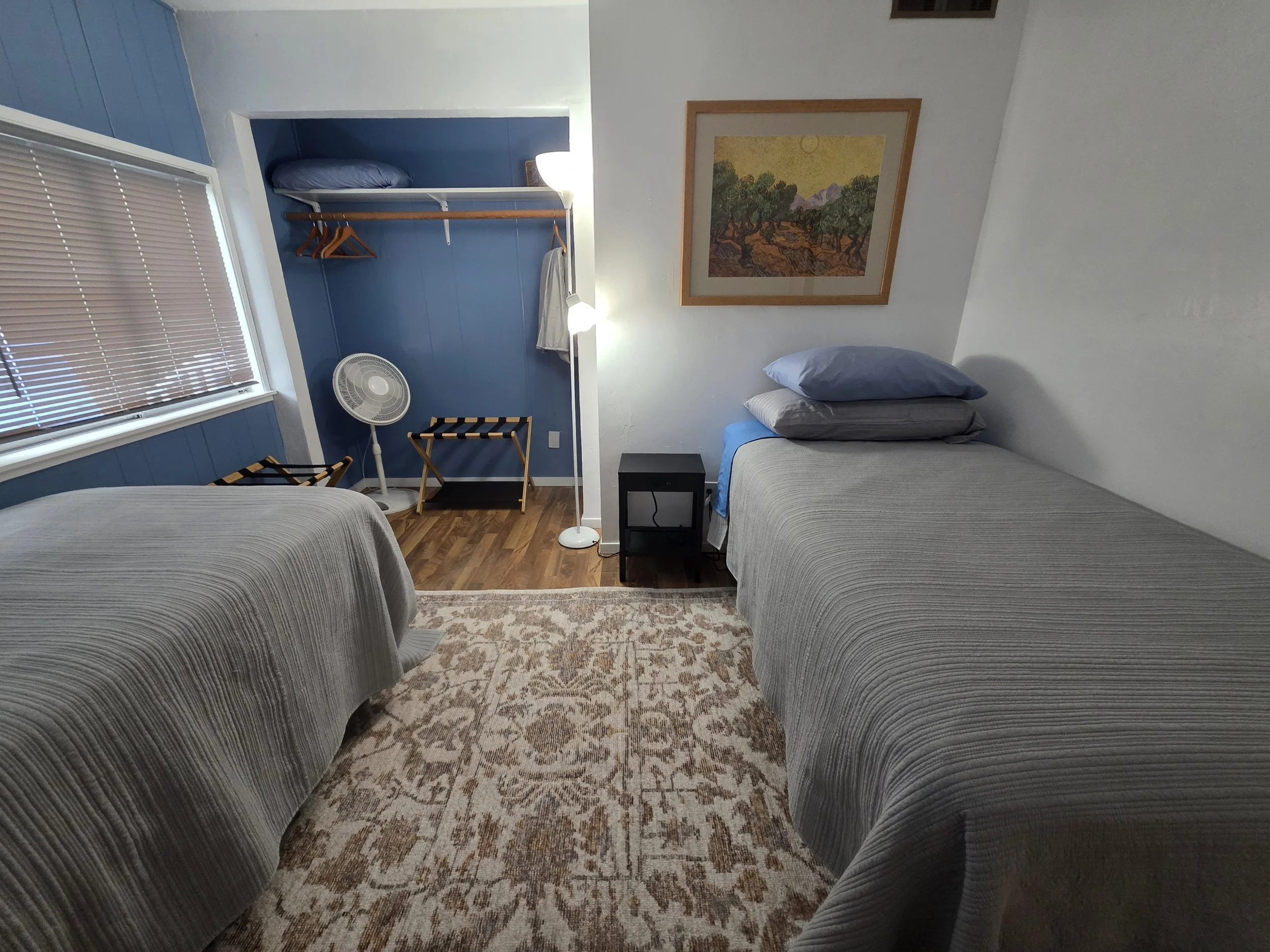 A bedroom with two beds, a window with blinds, a framed picture on the wall, a small black table between beds, a standing fan, a floor lamp, and an open closet with a blue accent wall.