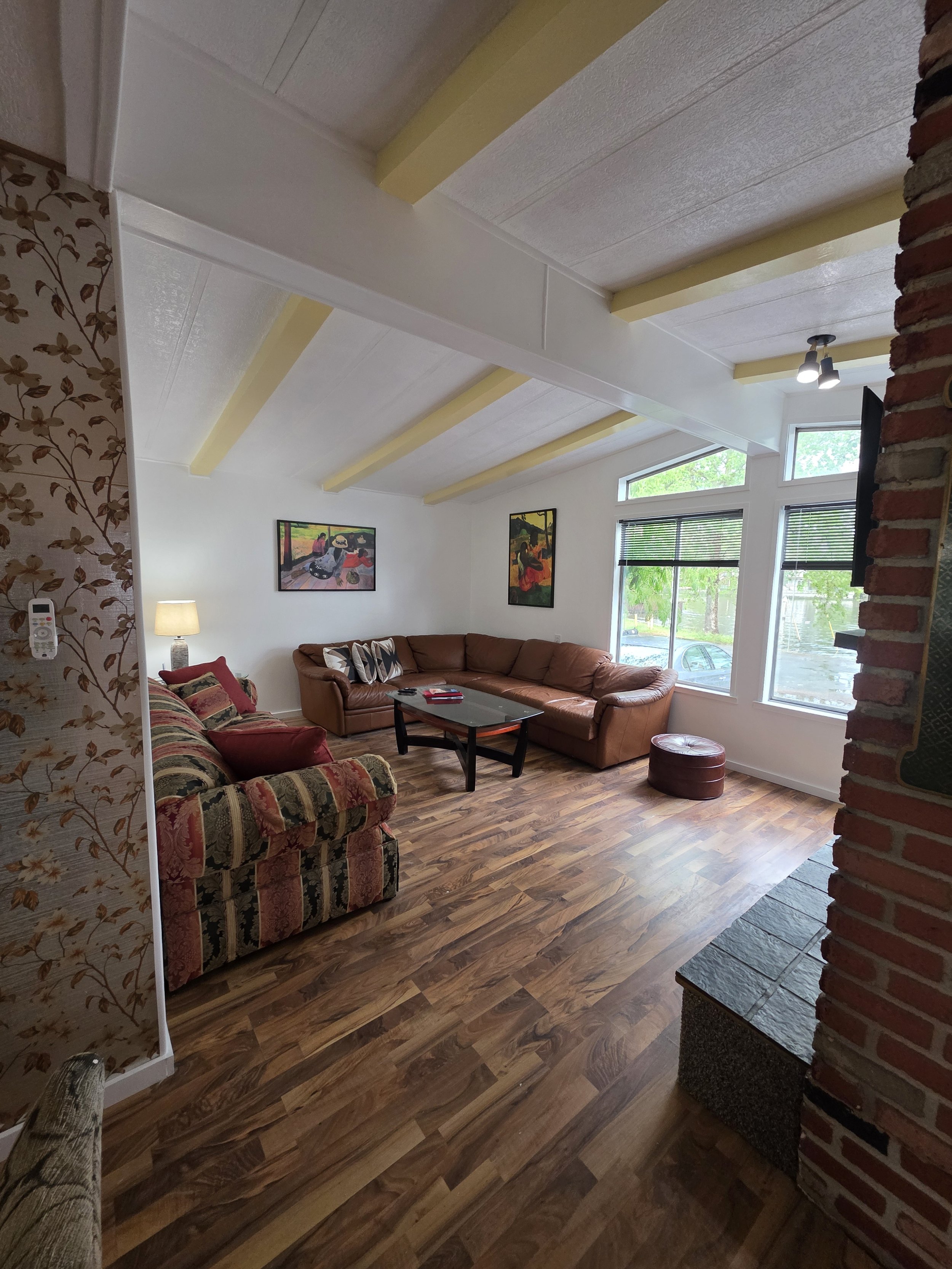 Living room with a brown sectional sofa, a floral patterned armchair, a glass coffee table, and wall art. Large windows with blinds let in natural light, and there is a brick fireplace on the right.