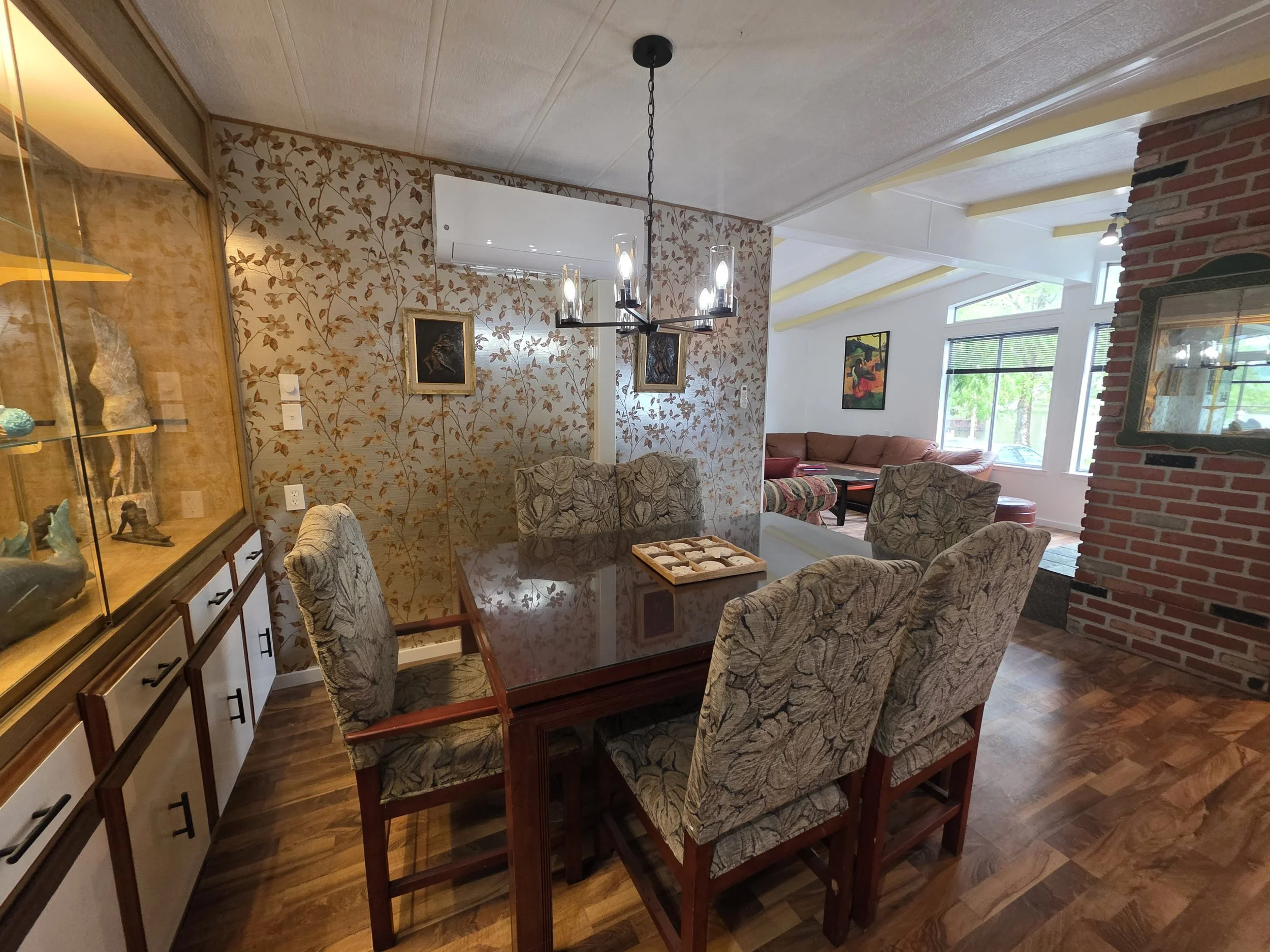 A dining room with a wooden table surrounded by upholstered chairs with floral fabric. A glass display cabinet with decorative items is on the left. The wall behind the table has patterned wallpaper with leaves, and two framed paintings hang on it. A