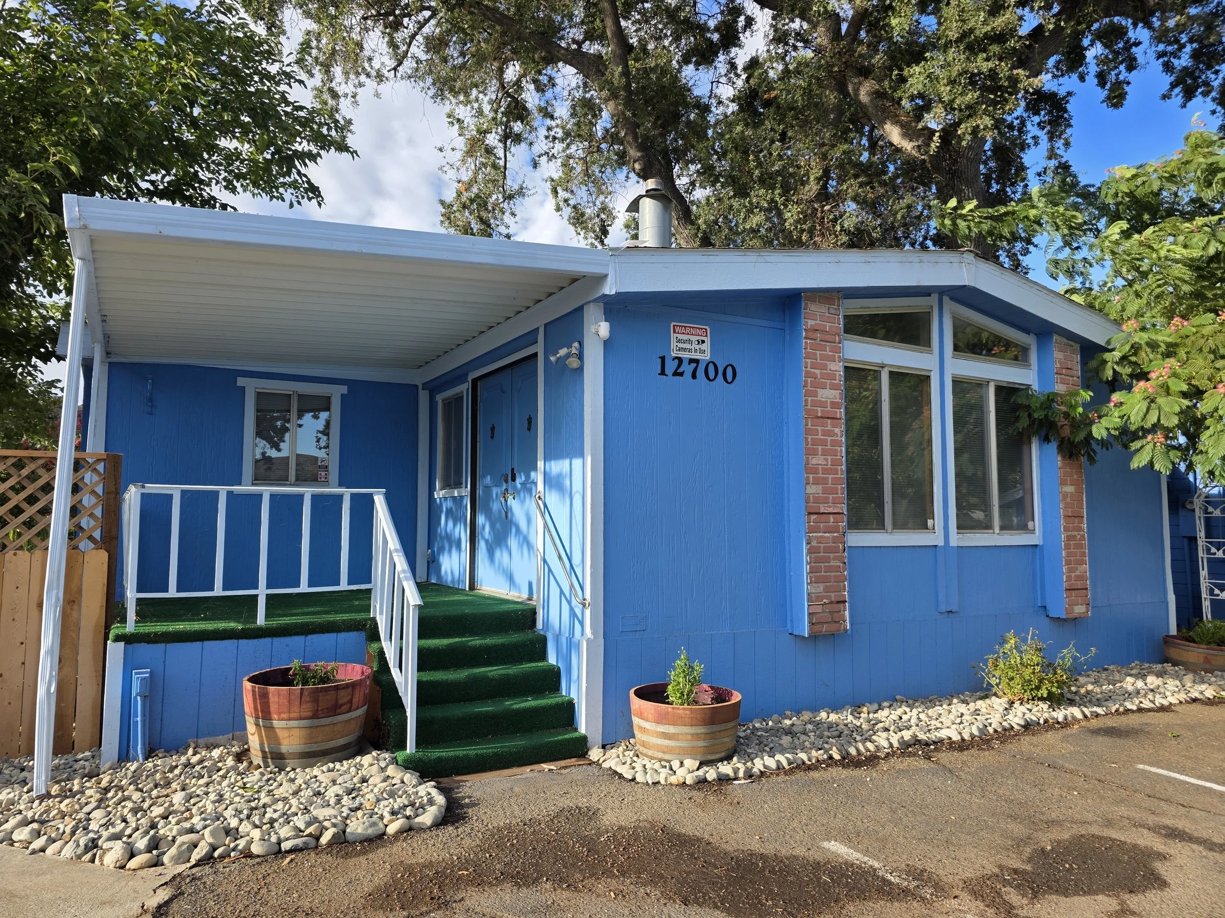Blue house with white trim, brick accents, and an entry porch with green carpeting, front steps, potted plants, and a parking lot.