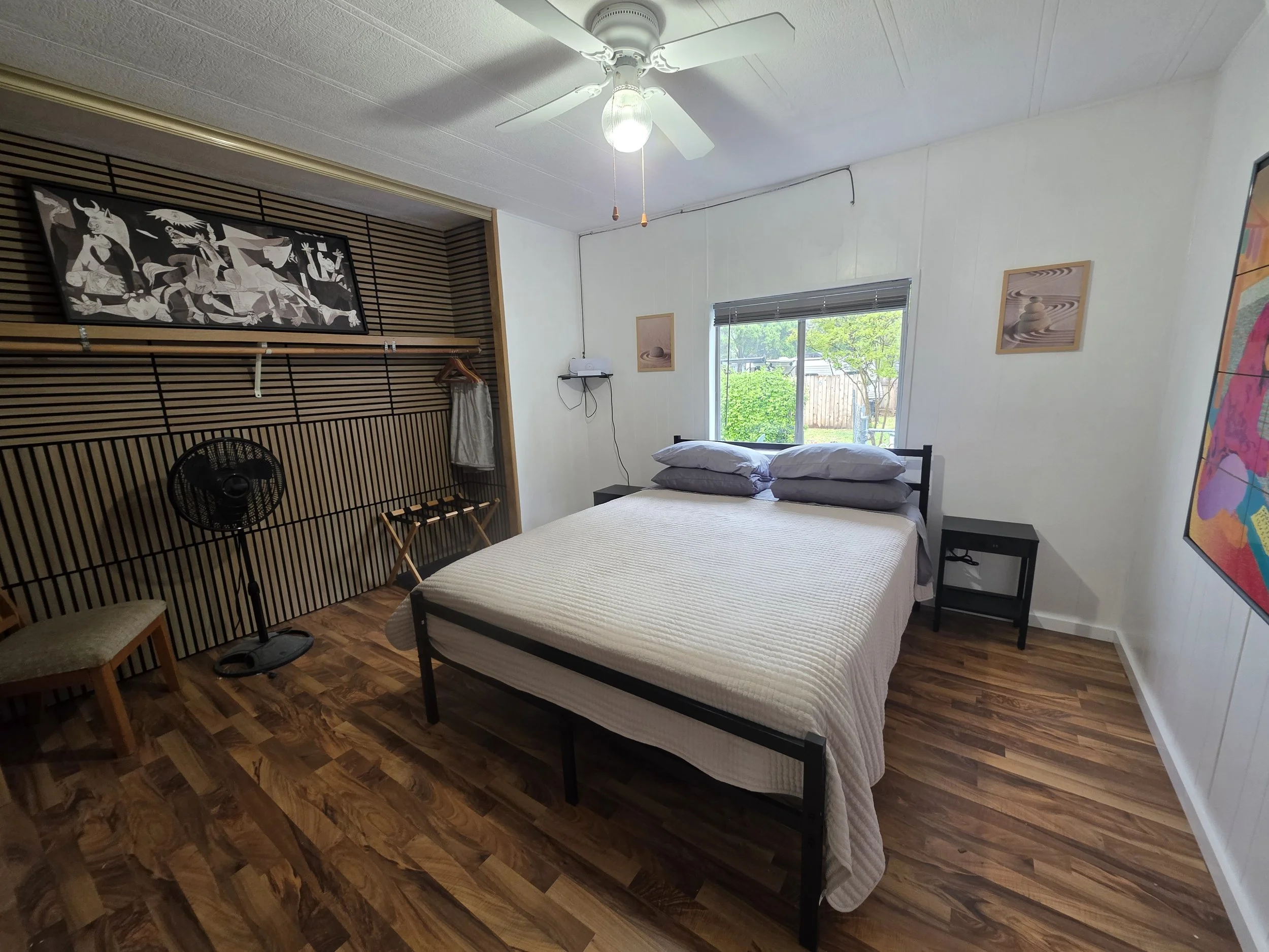 A bedroom with a bed, two nightstands, and artwork on the walls. There are two pillows on the bed, and a window showing a view of greenery outside. An open closet with a fan and hanging rod is visible, as well as a ceiling fan with a light. The floor